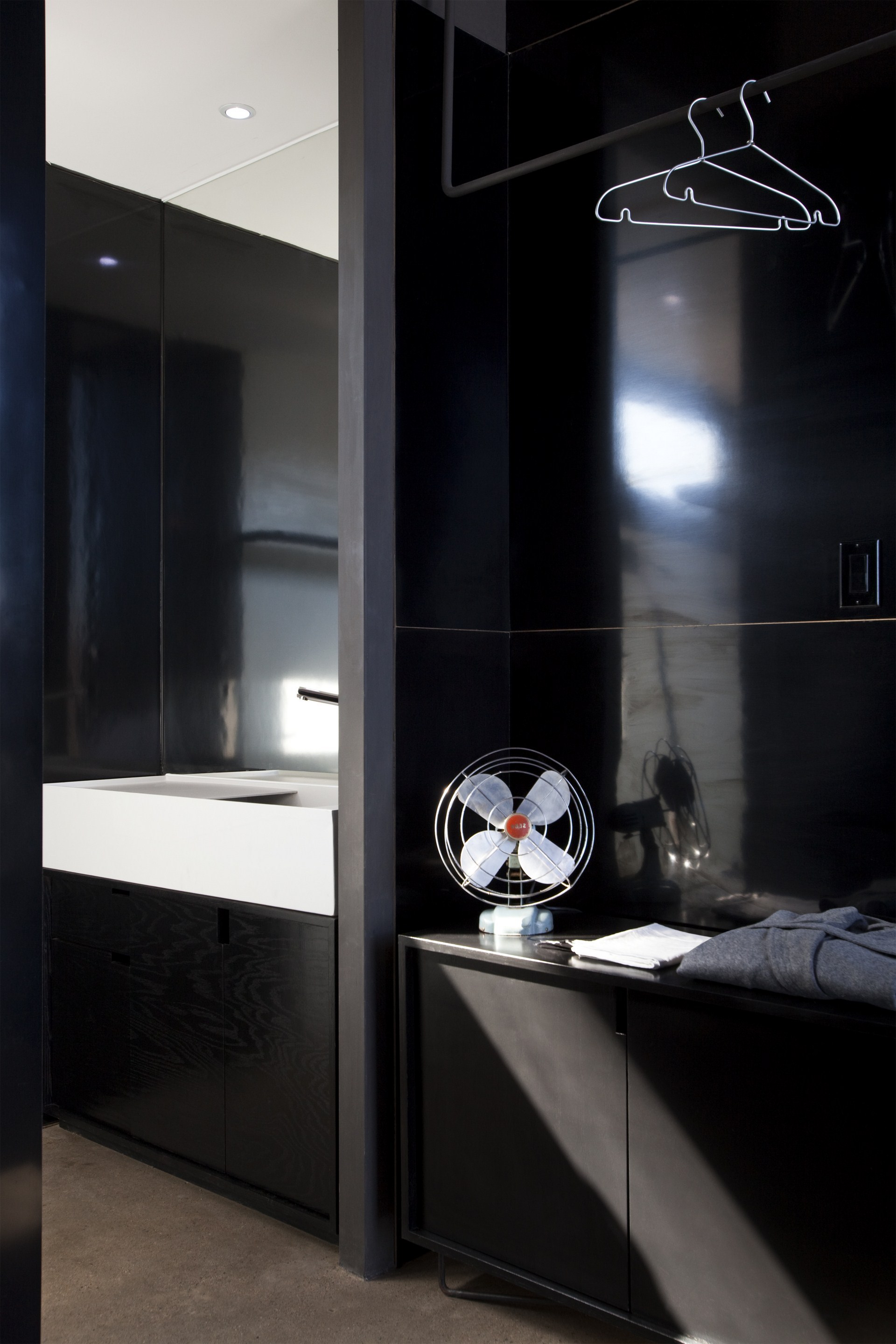 Black lacquered cabinetry with a vintage desk fan, wire hangers and a white basin, lit by a window reflection