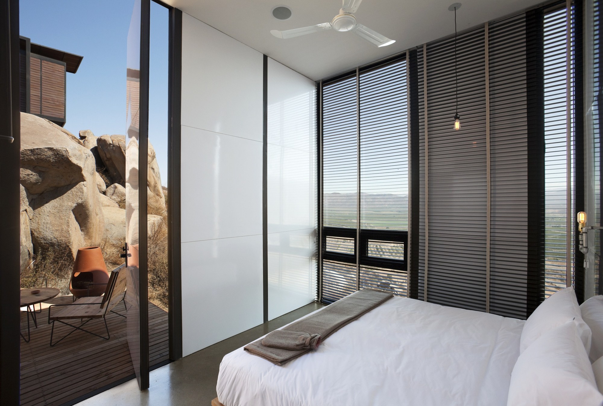 Bedroom corner with a pivoting glass door open to a timber deck and boulders under clear blue sky