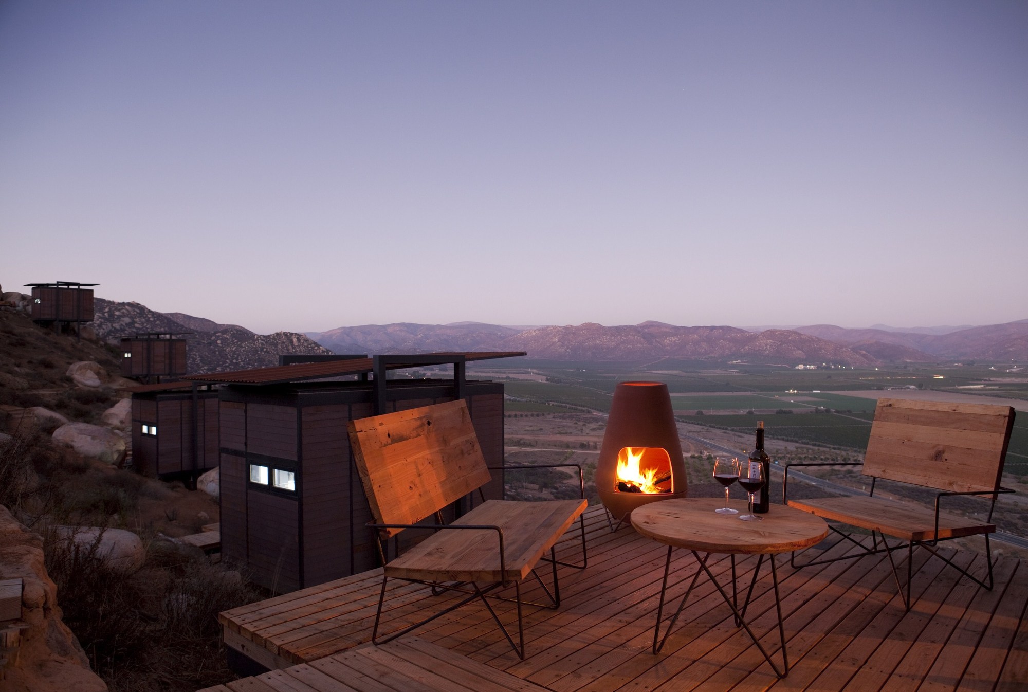 Timber deck with a clay fireplace, wood benches and wine glasses facing a valley at dusk