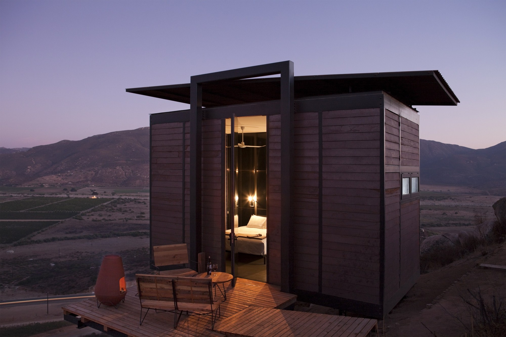 Timber-clad cabin with a cantilevered roof and glowing bedroom seen through an open door at dusk