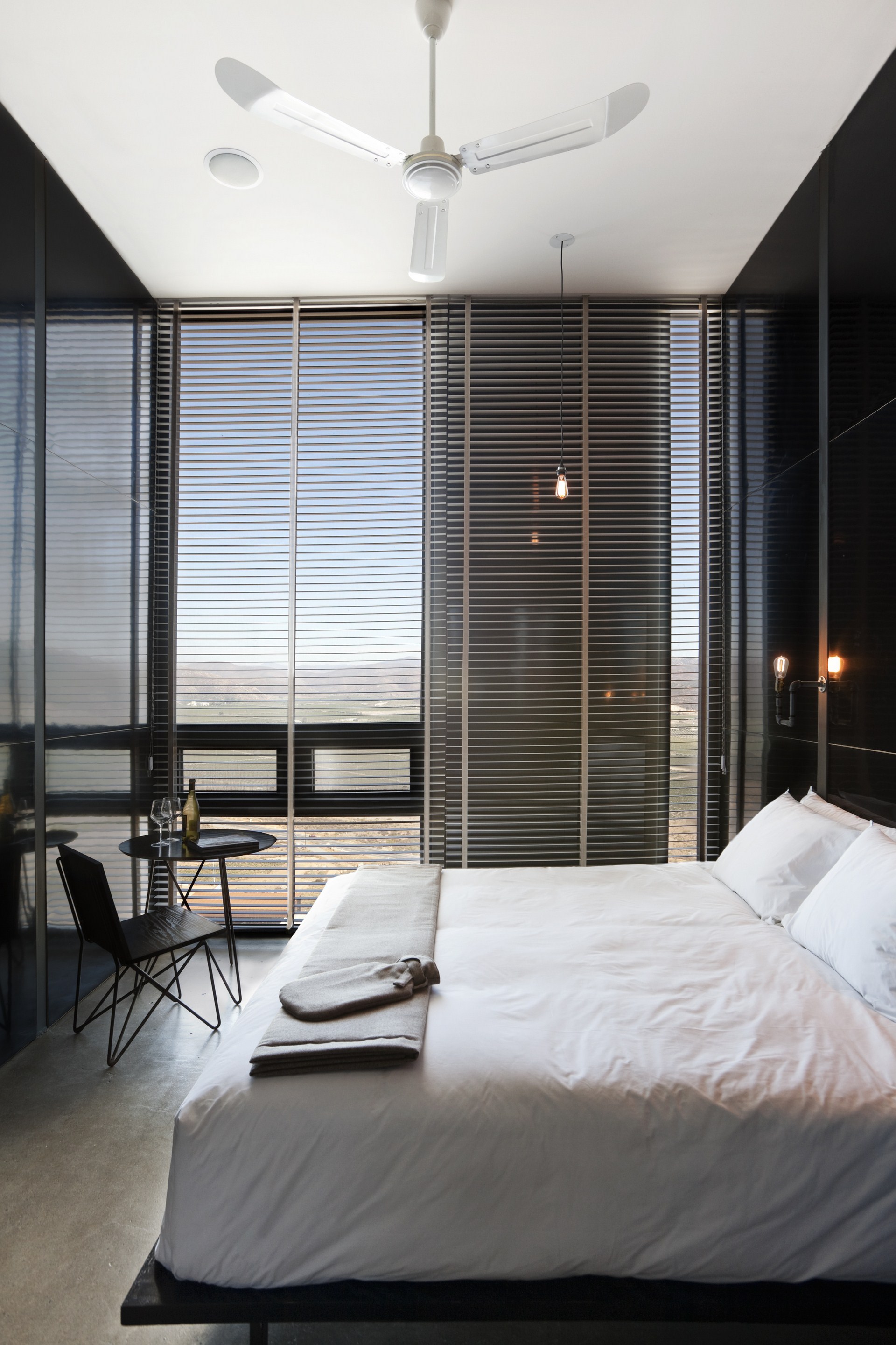 Bed with folded grey blanket facing full-height slatted blinds, a pendant bulb and a small bistro table beside it