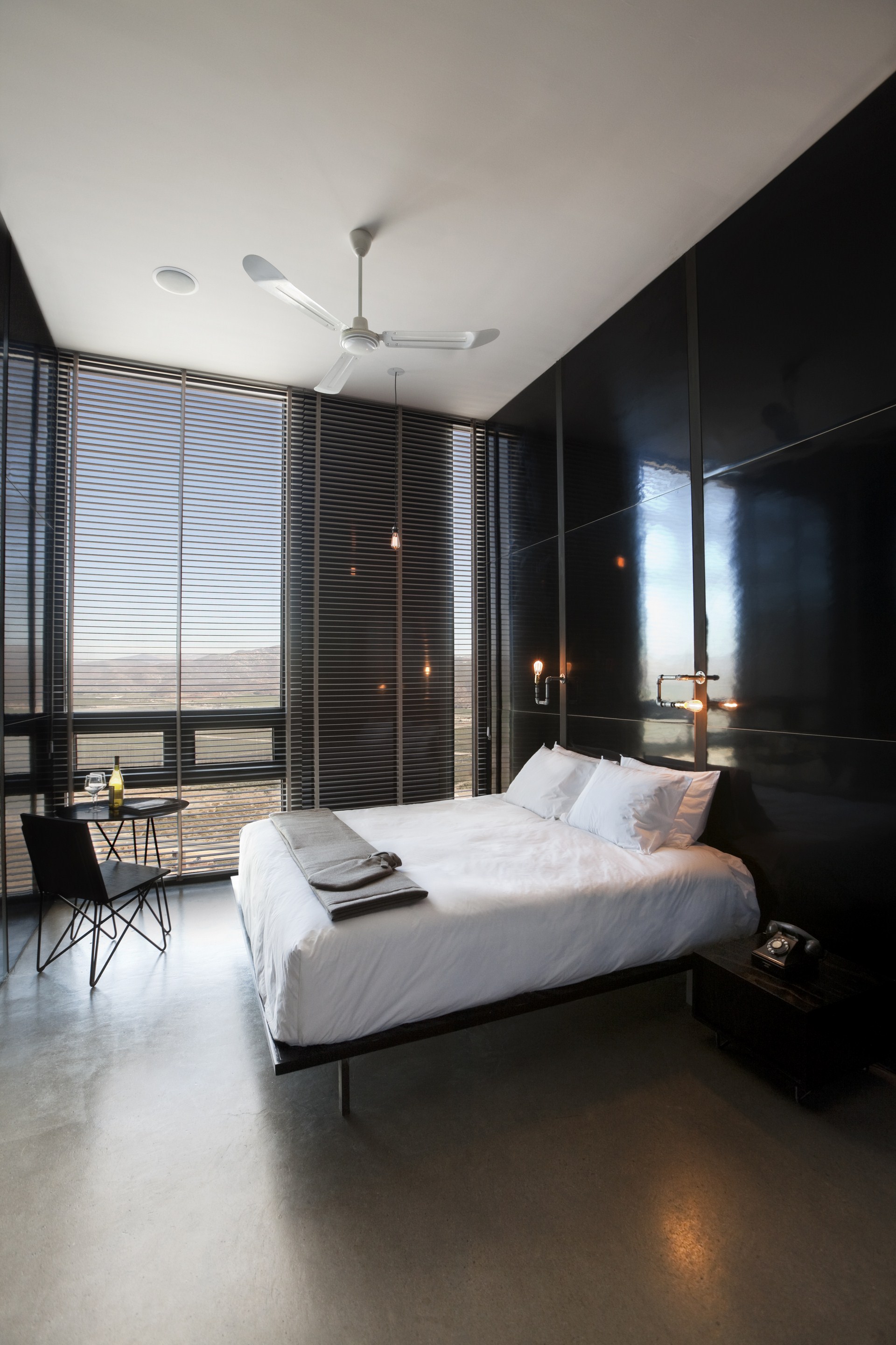 Bedroom with white bed against a glossy black panel wall, ceiling fan above and slatted blinds filtering desert hillside light