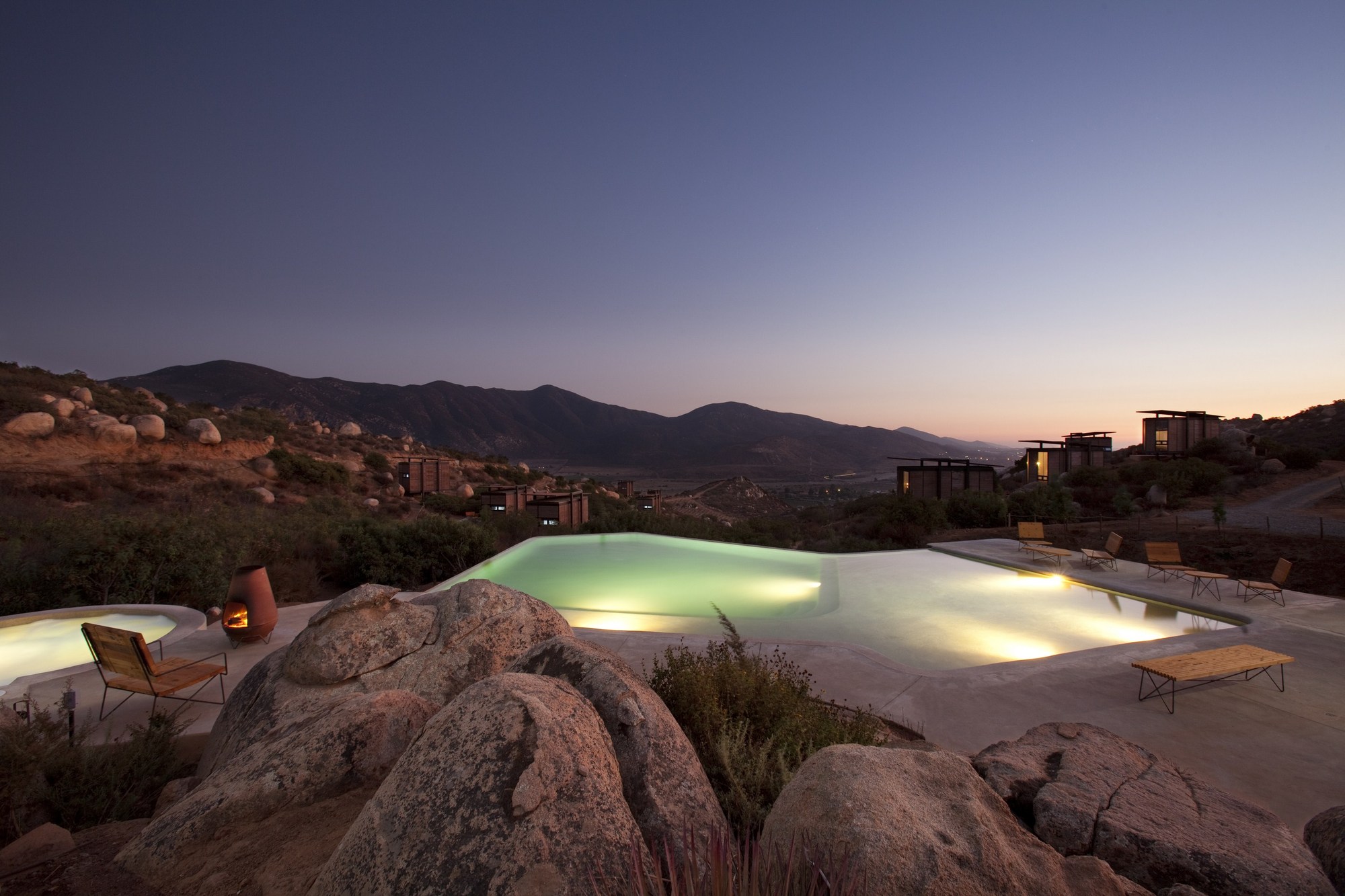 Infinity pool glowing among granite boulders at dusk, with small cabins scattered on the hillside beneath a fading purple sky