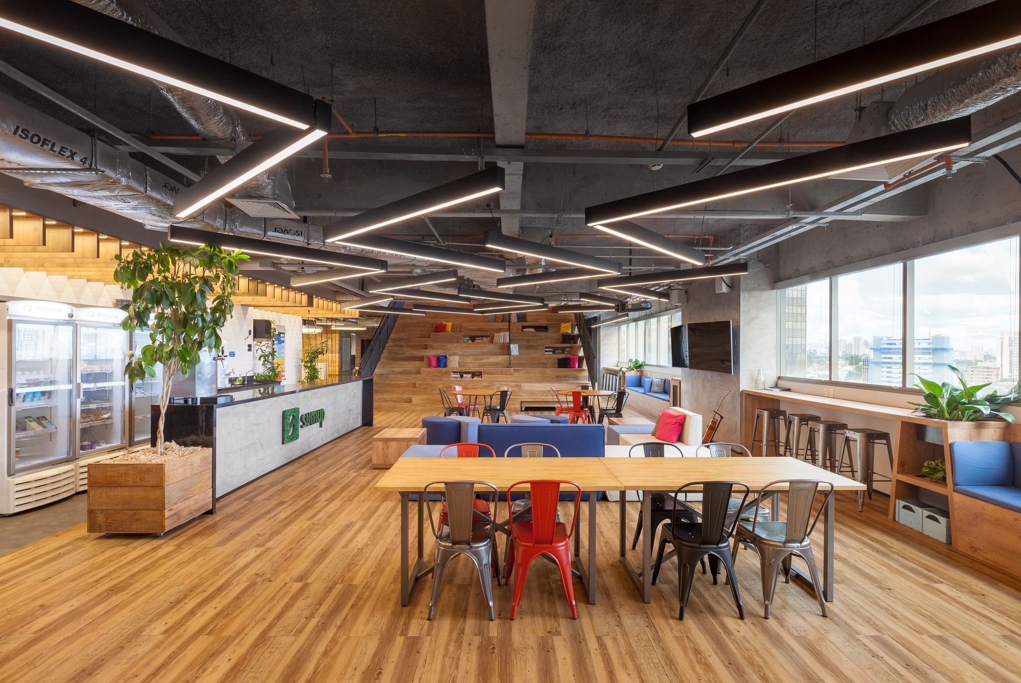 Long communal table with red and grey metal chairs beside a reception counter, under linear pendant lights and exposed ducts