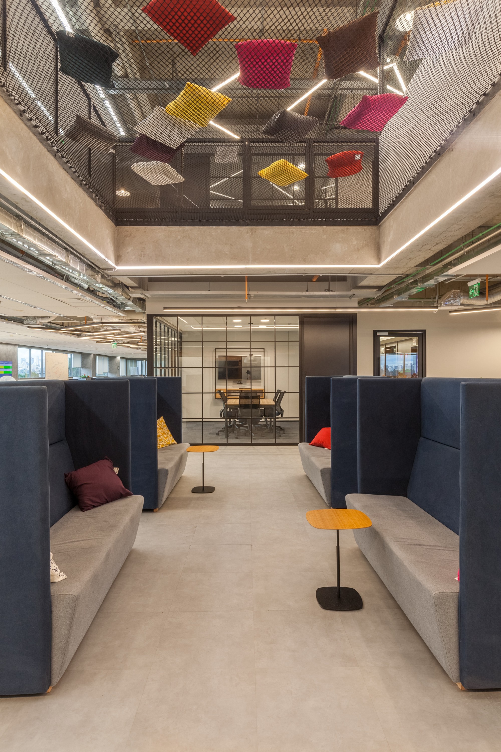 Navy high-back booth sofas facing a glass-walled meeting room, with colorful cushions hanging in a net above
