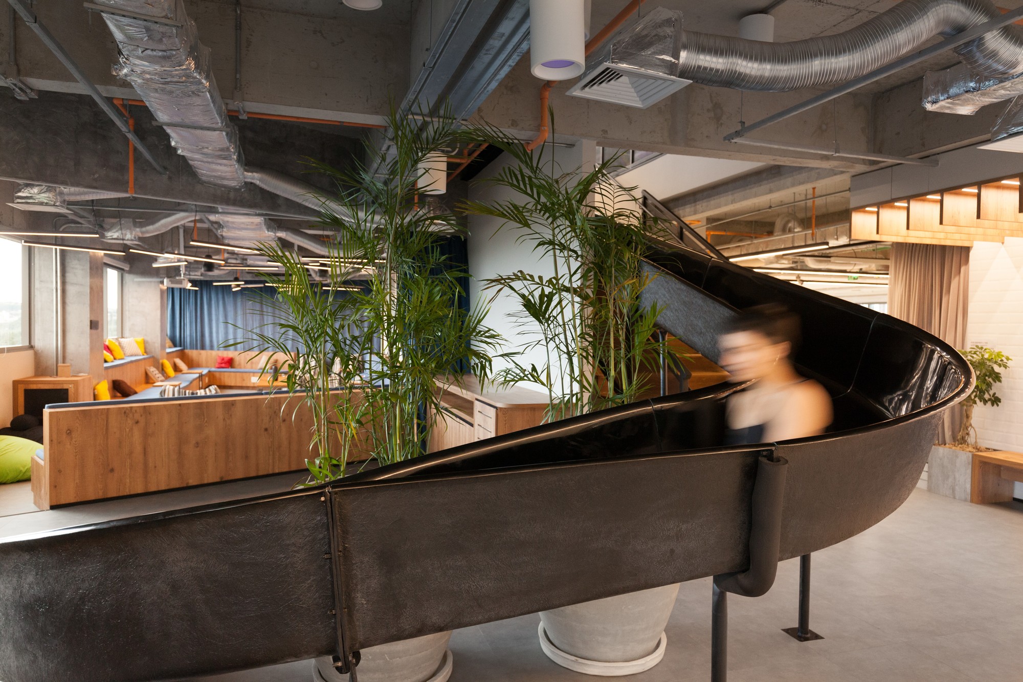 Black curved slide sweeping past potted palms, with a blurred person sliding down in soft daylight