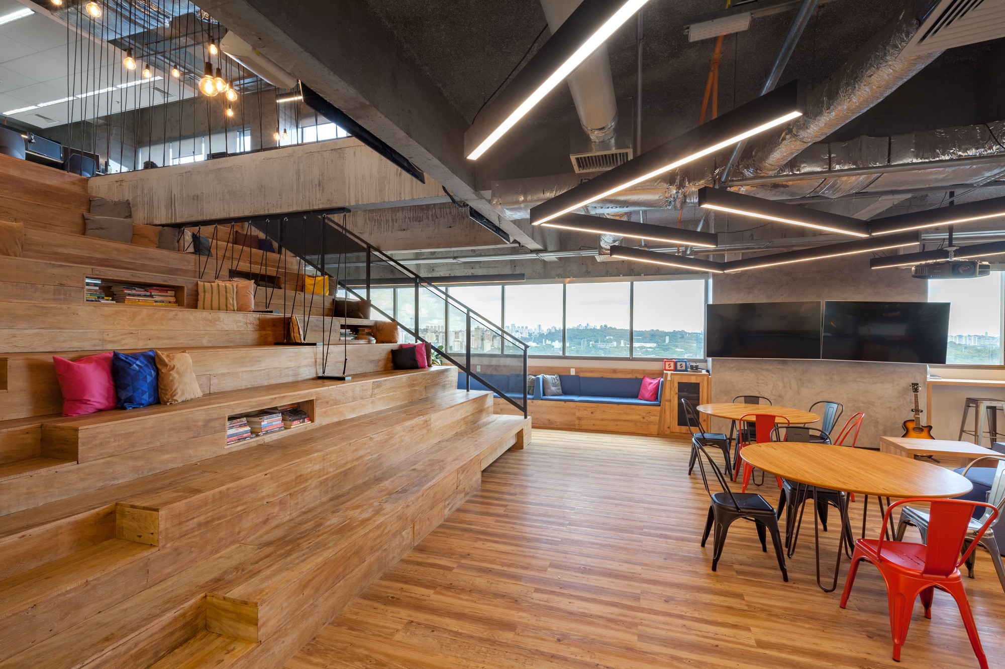 Timber stepped seating with books and bright cushions beside round tables, with wide windows overlooking the skyline in daylight