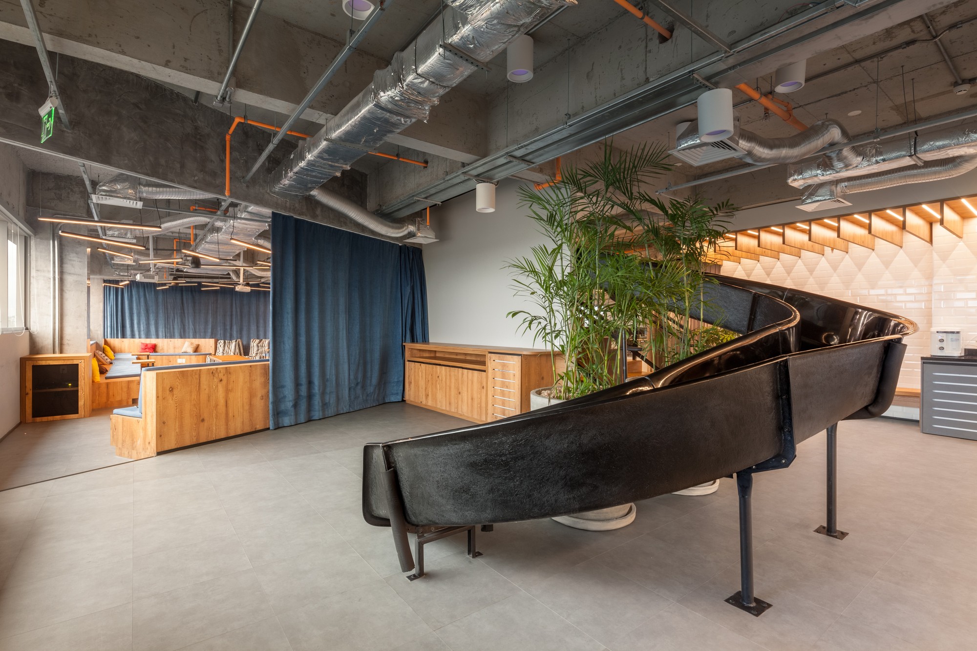 Black curved fiberglass slide sweeping past a potted palm, with a wood-paneled lounge and blue curtain behind under exposed ducts