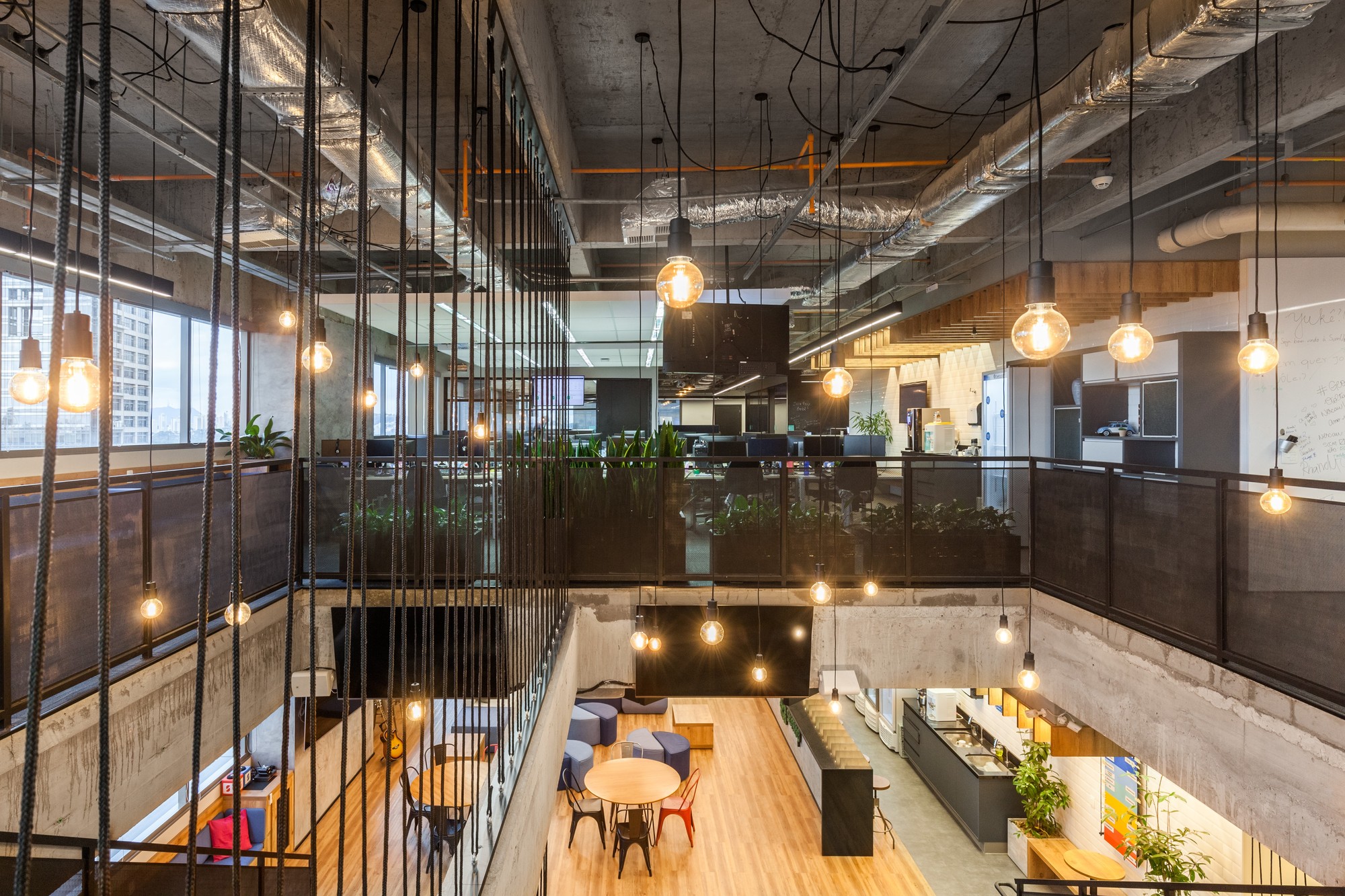 View from an upper walkway down into a double-height office atrium, with bare bulb pendants hanging on cords over planters and a wood floor