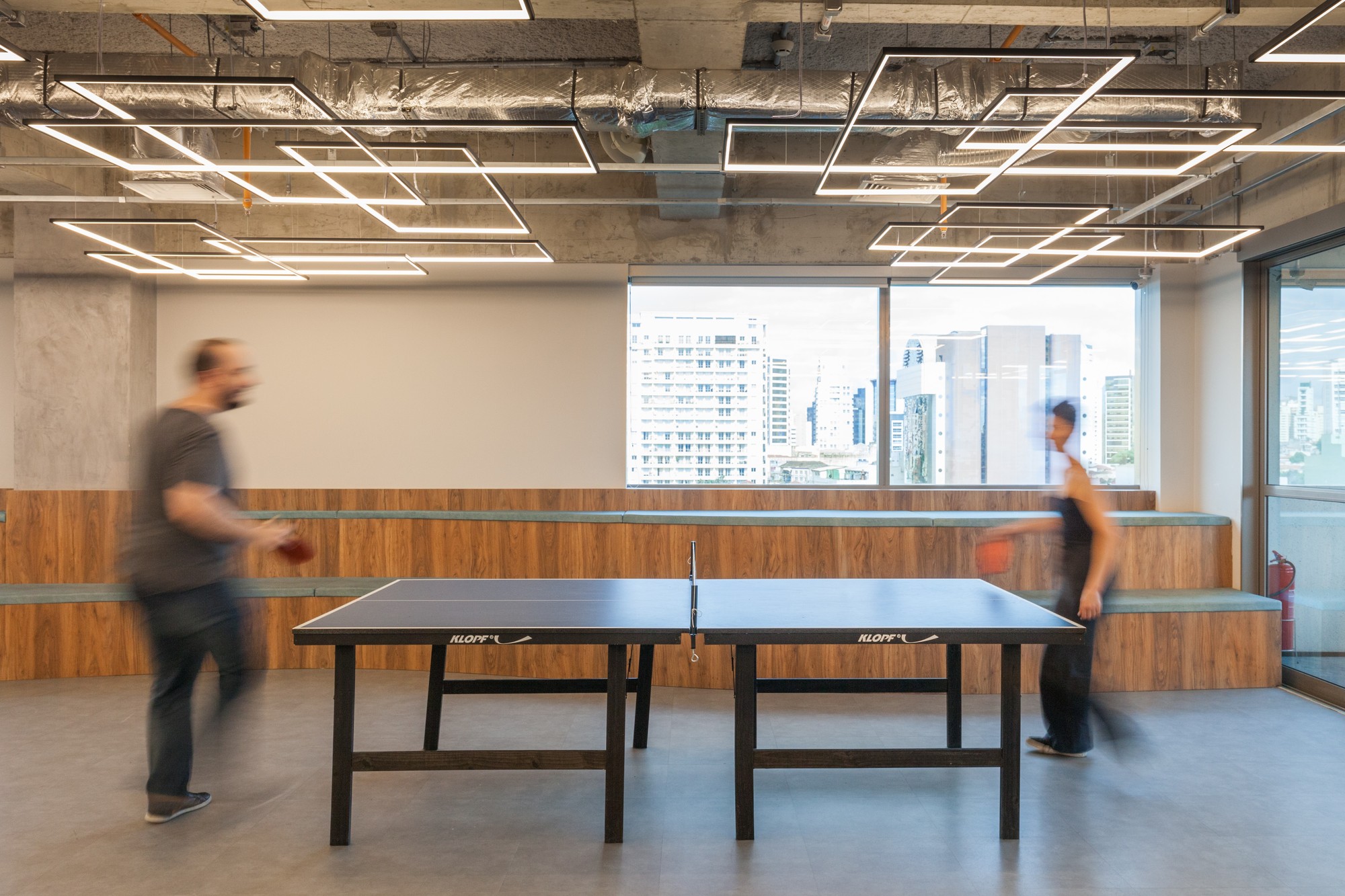 Two blurred players at a ping-pong table beneath rectangular linear light fixtures, with city towers visible through the window behind