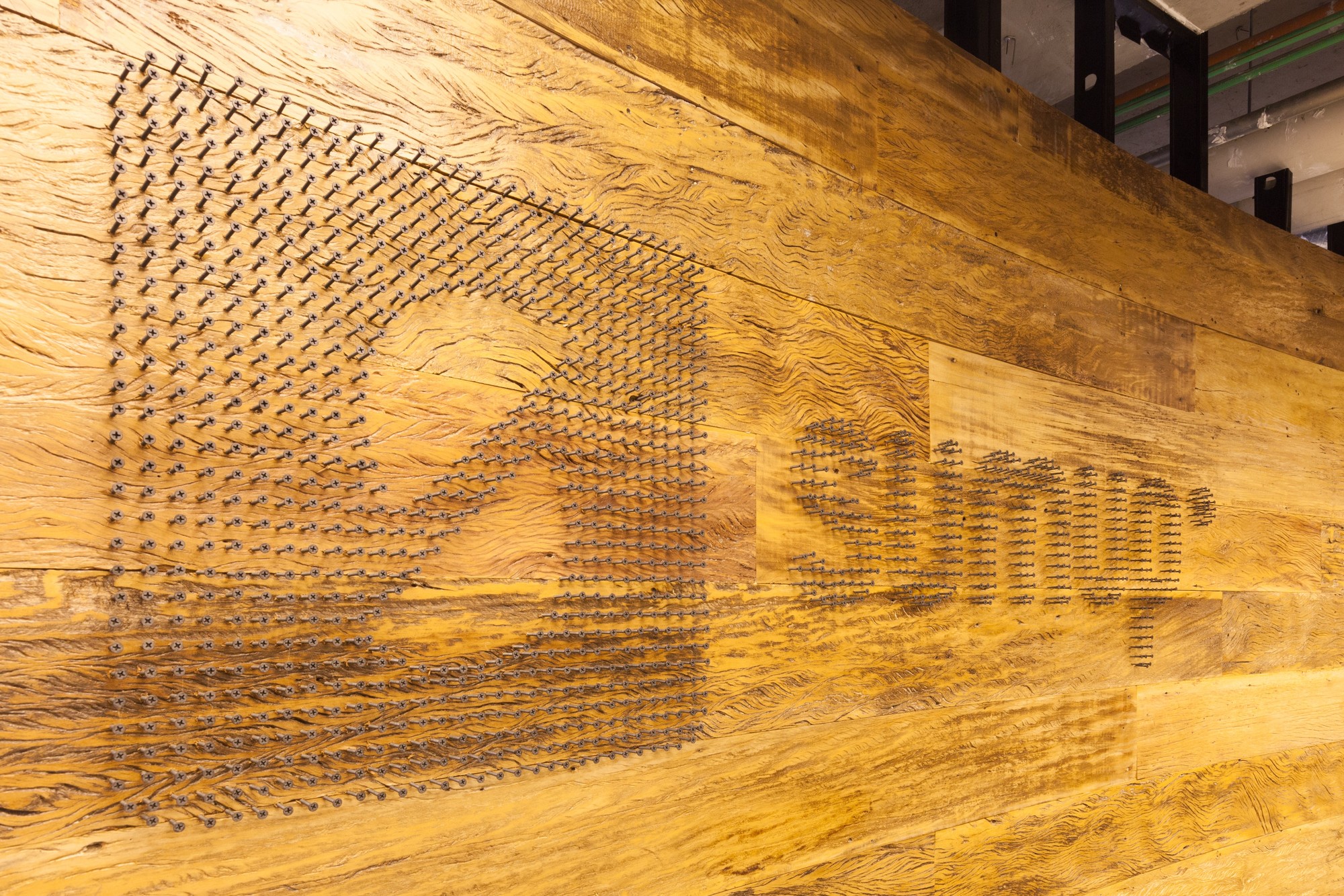 Close-up of a reclaimed wood wall with a logo and lettering formed from rows of protruding nails, lit from the side