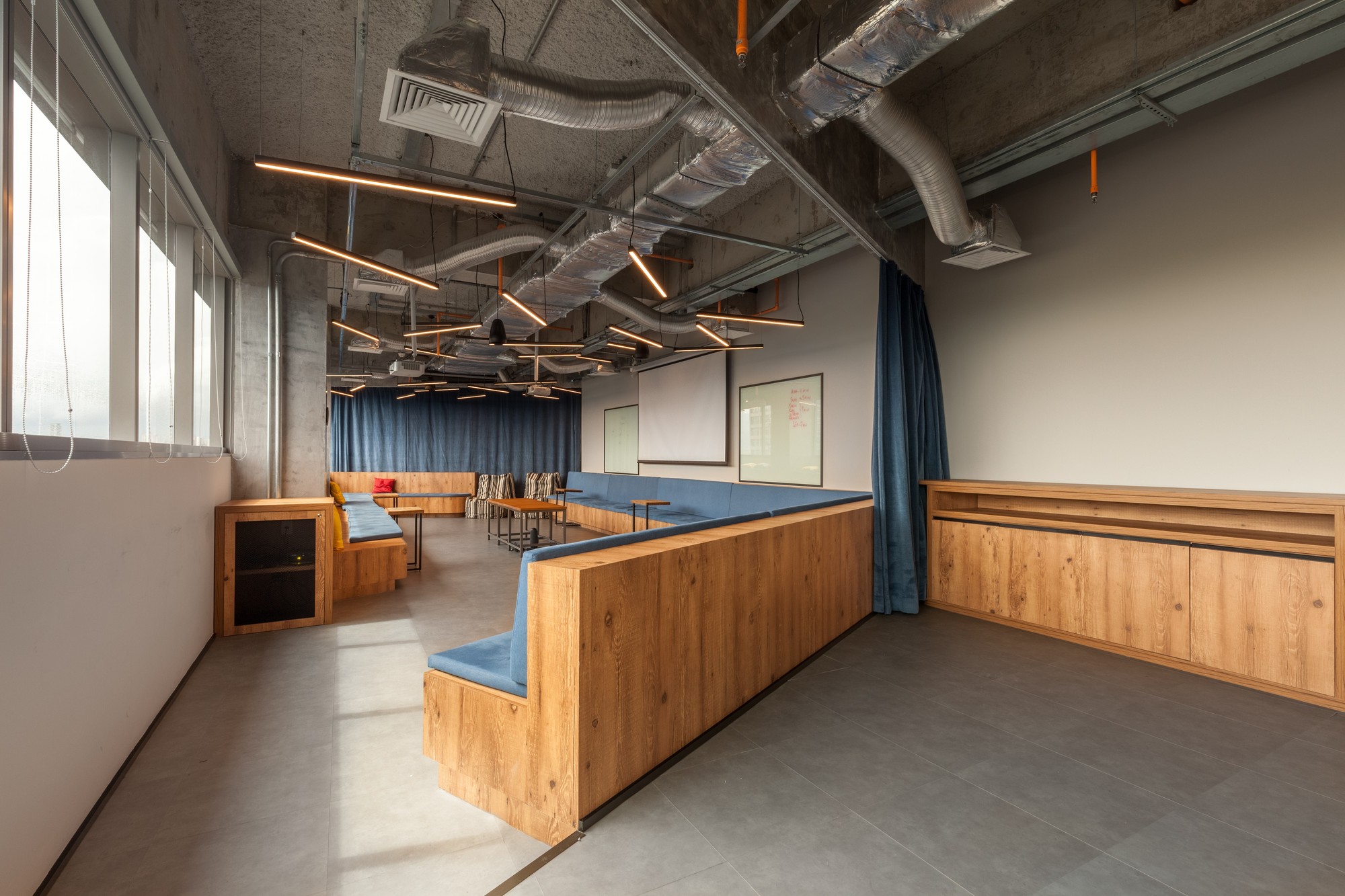 Meeting room with a wood-clad built-in bench with blue cushions, a projection screen, blue curtains and copper-toned pendant lights under exposed ductwork