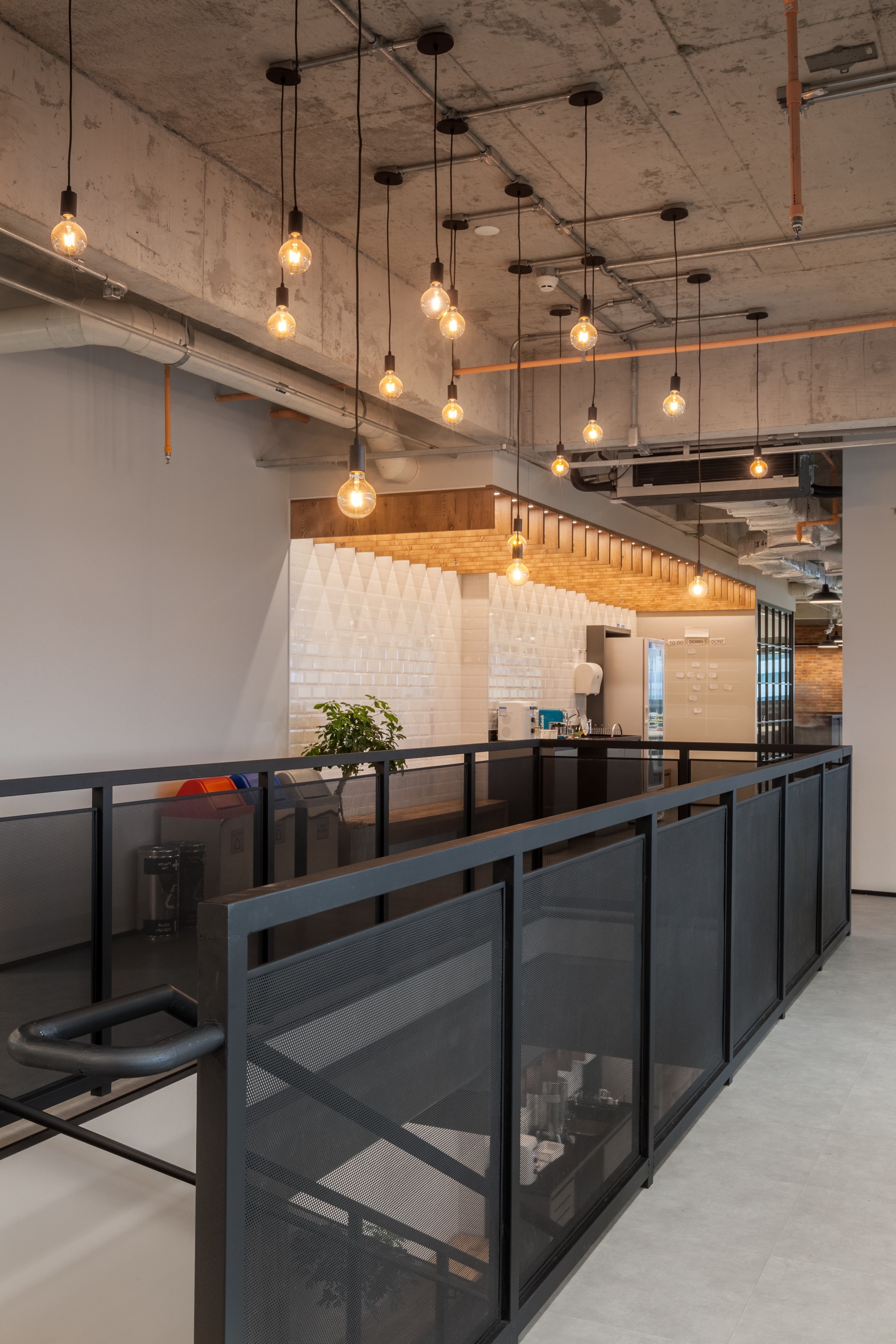 Black mesh-panel balustrade overlooking a white tiled kitchen nook, with bare filament bulbs hanging from an exposed concrete ceiling