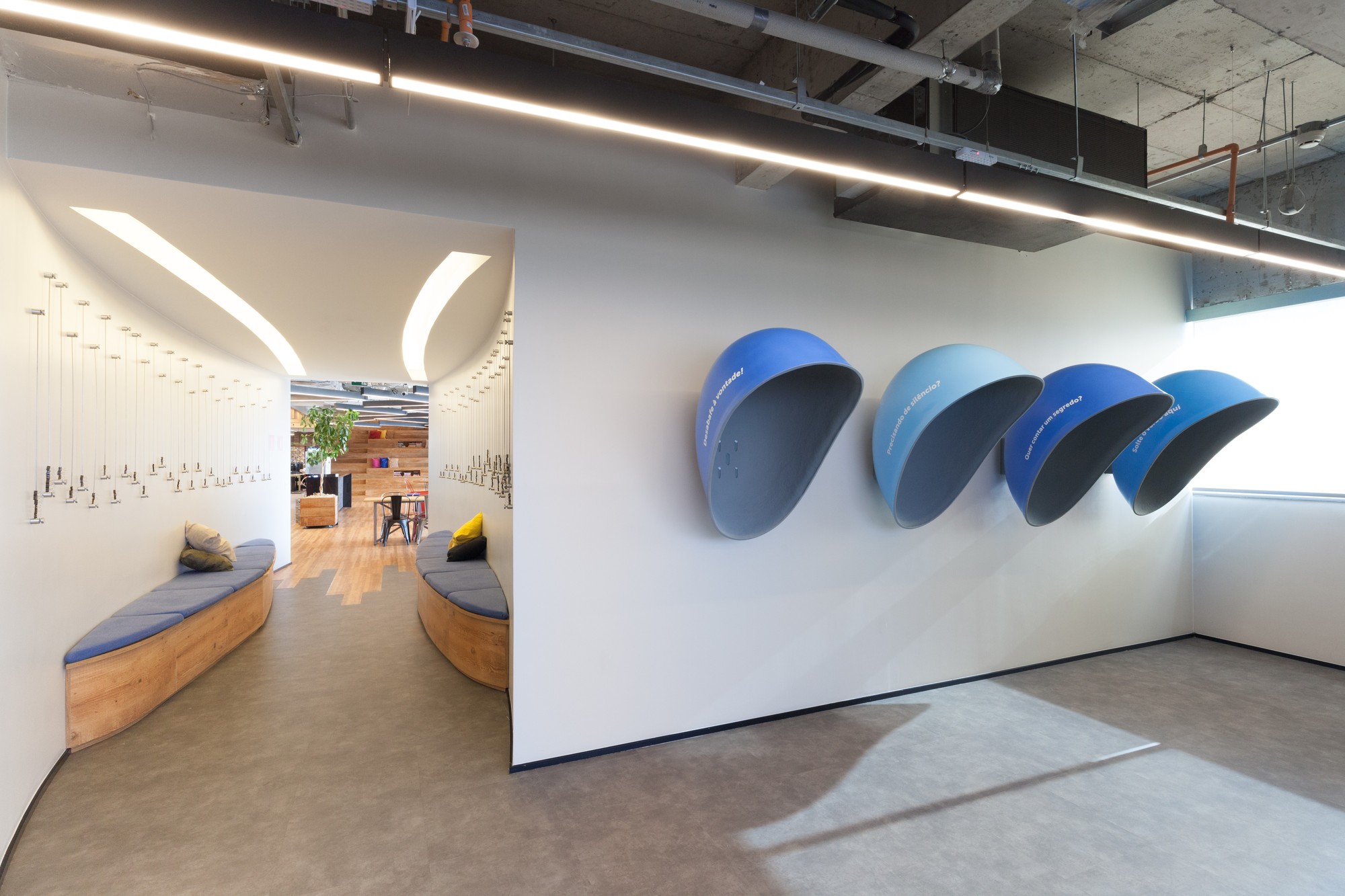 Row of blue wall-mounted phone pods beside a curved passage with wood benches, lit by soft ceiling slots and daylight