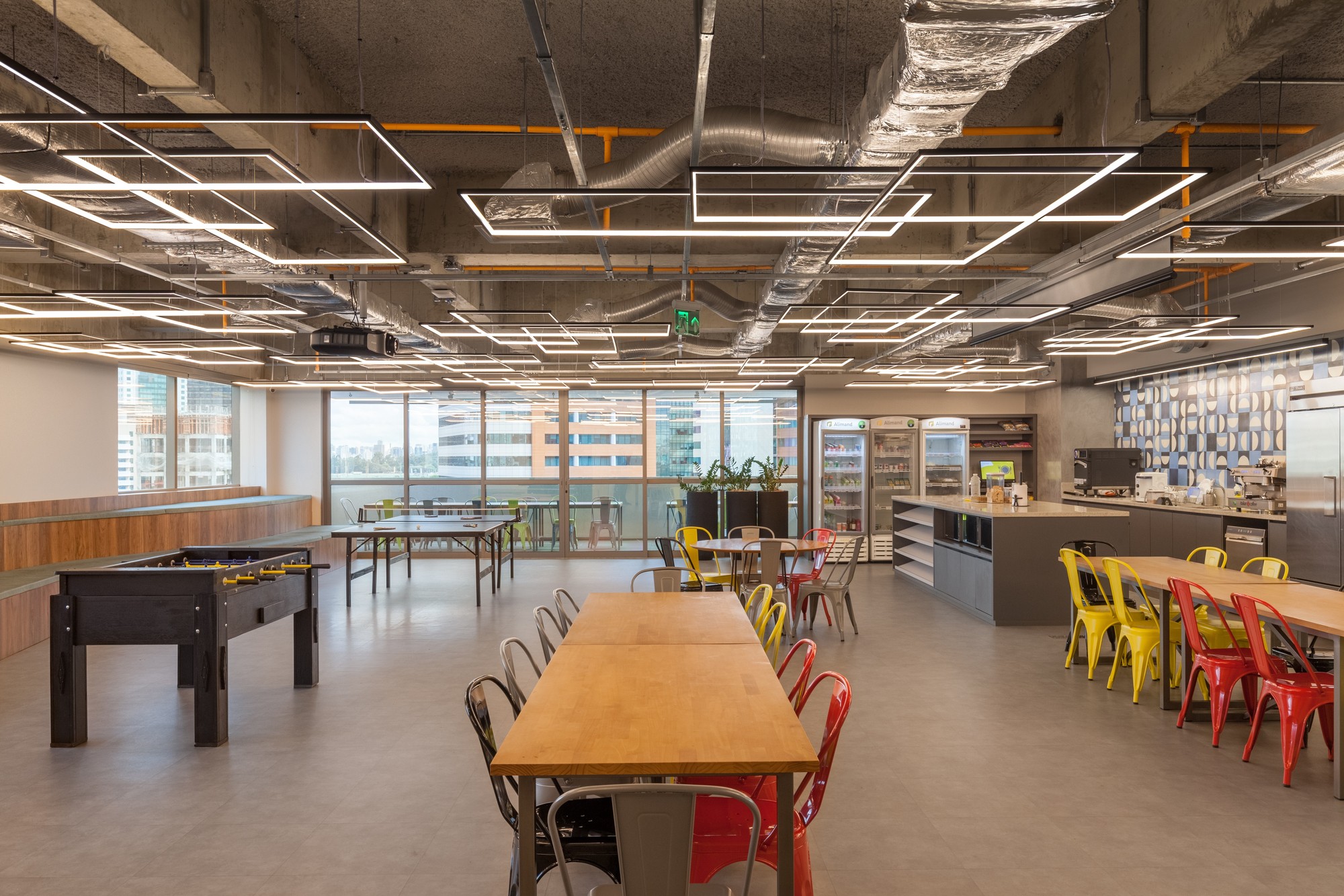 Open cafeteria with long timber tables, red and yellow metal chairs, a foosball table and linear light fixtures under exposed ducts