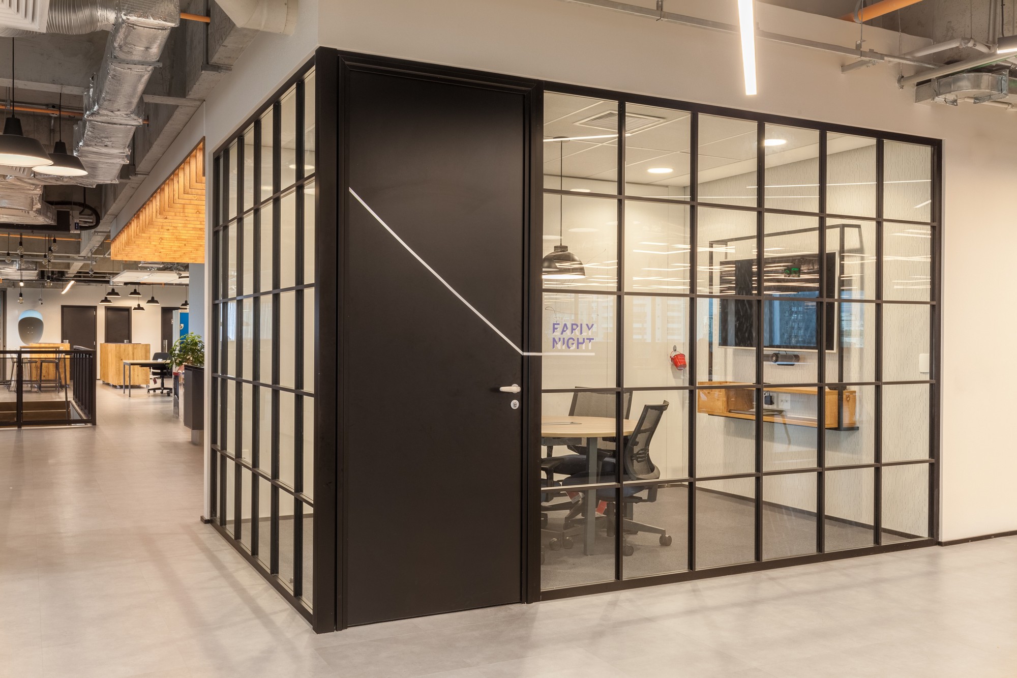 Black steel-framed glass meeting room on a polished concrete floor, with a corridor of pendant lamps stretching behind it