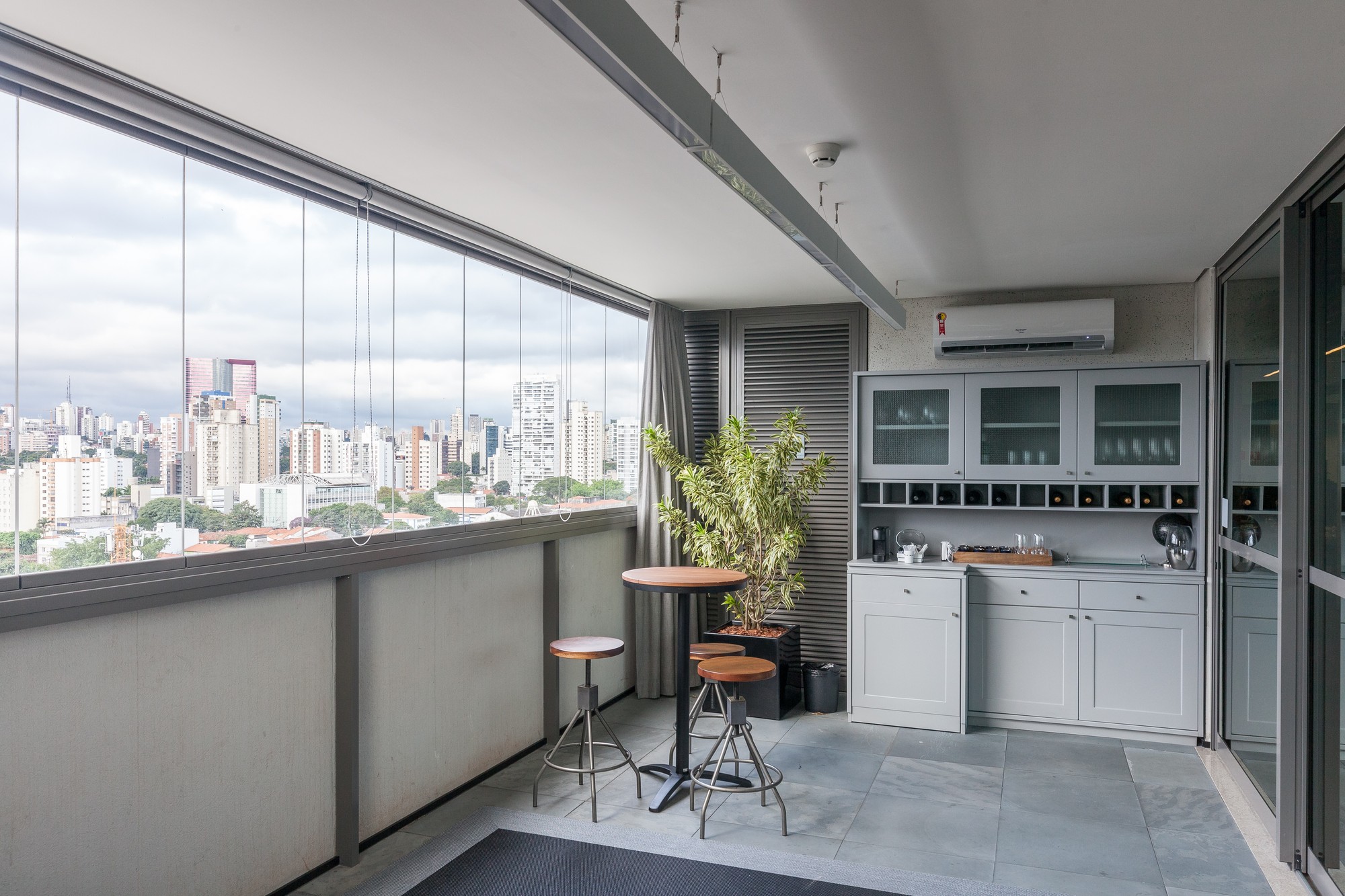 Glazed balcony with a grey cabinet, round bar table and stools, looking out over a hazy city skyline under cloud