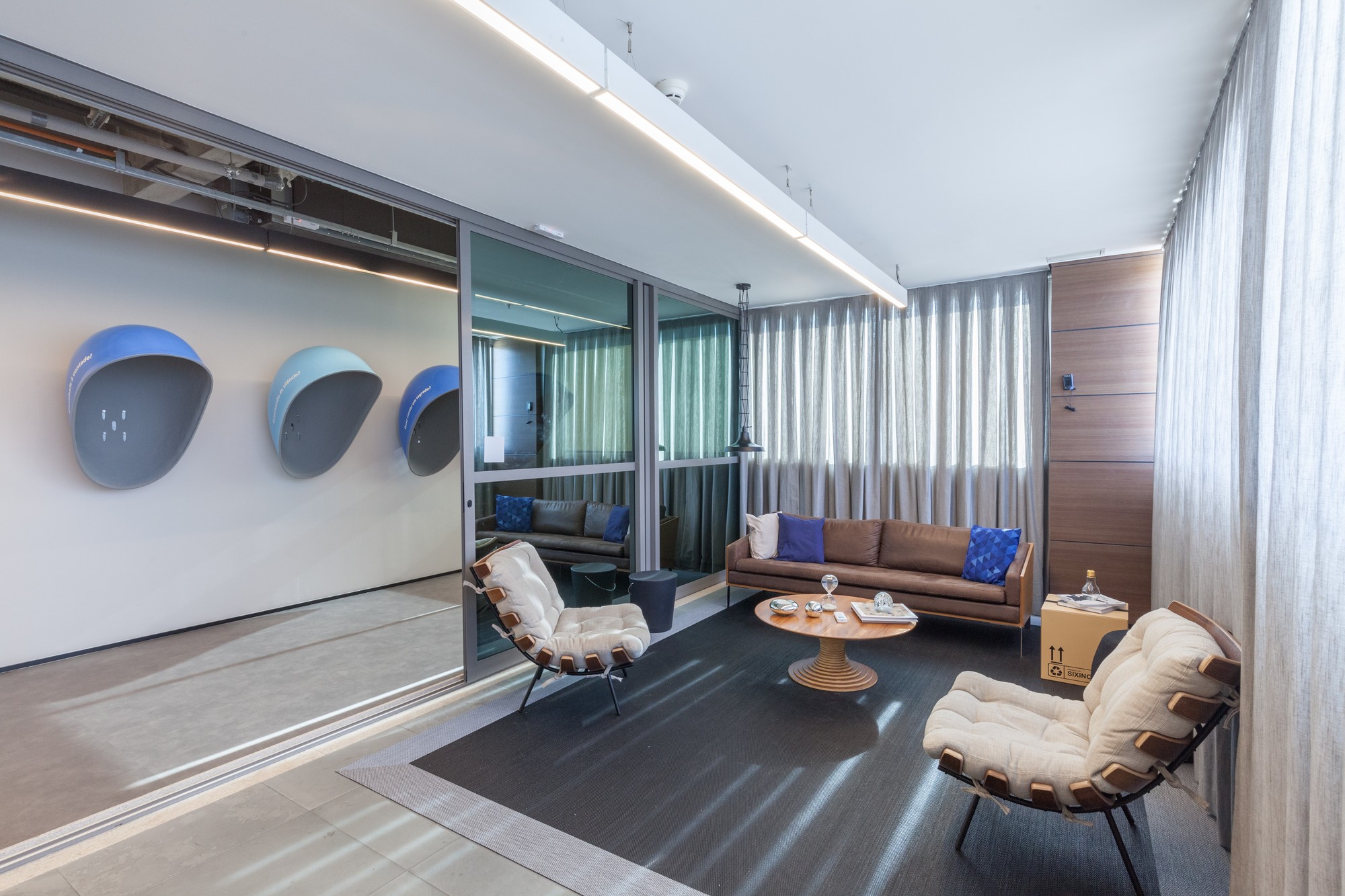 Same lounge with the glass partition slid open, revealing blue phone-booth shells on a white wall and sunlight striping the dark rug