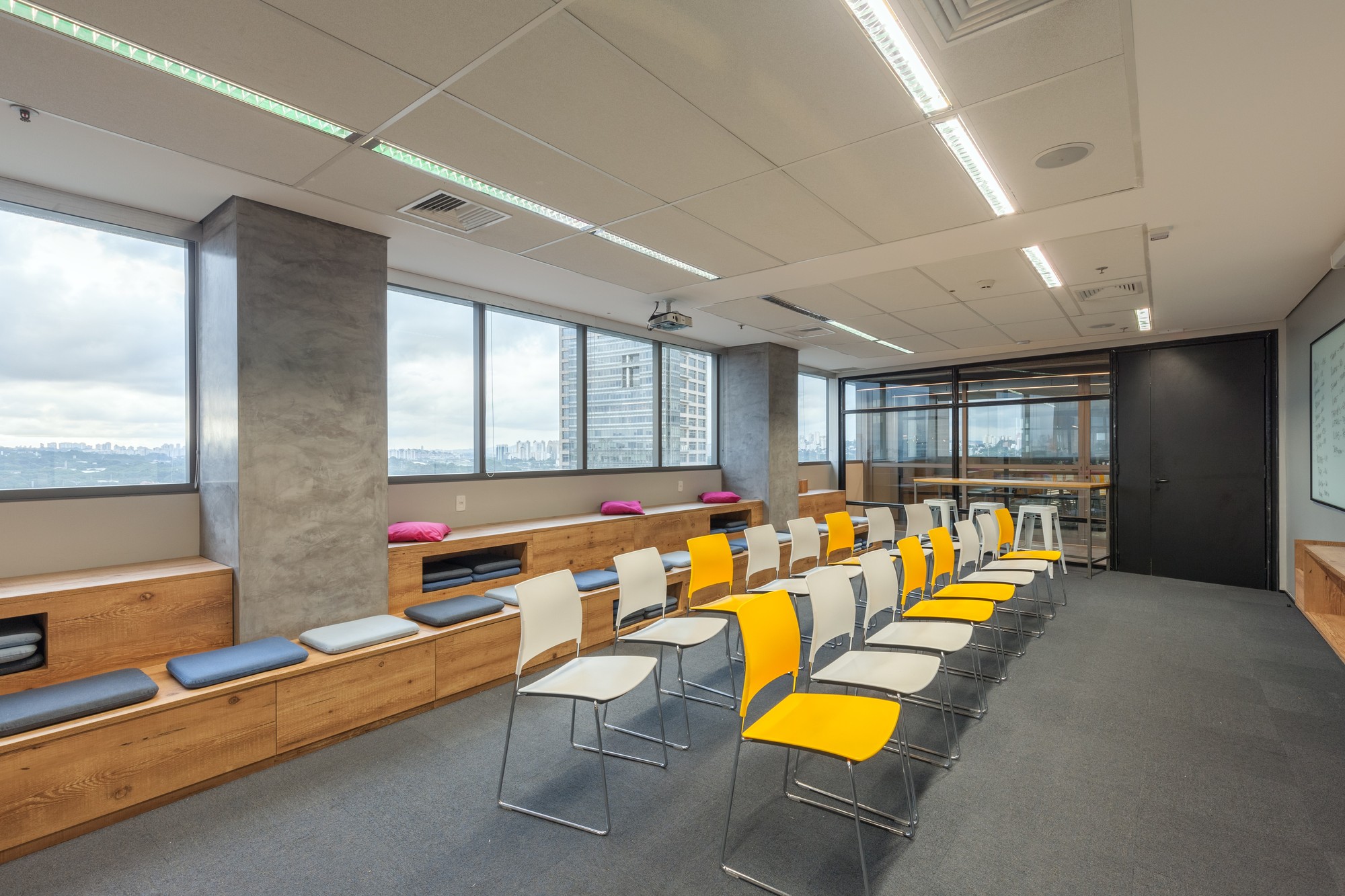 Training room with rows of yellow and white chairs facing built-in timber bench seating, concrete columns and a distant city skyline through the windows