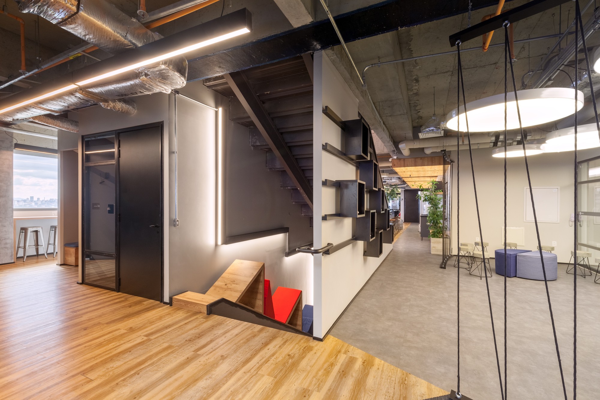 Open stair descending beside a wall of black shelving boxes, with exposed ducts and round pendant lights overhead in a concrete-ceilinged office