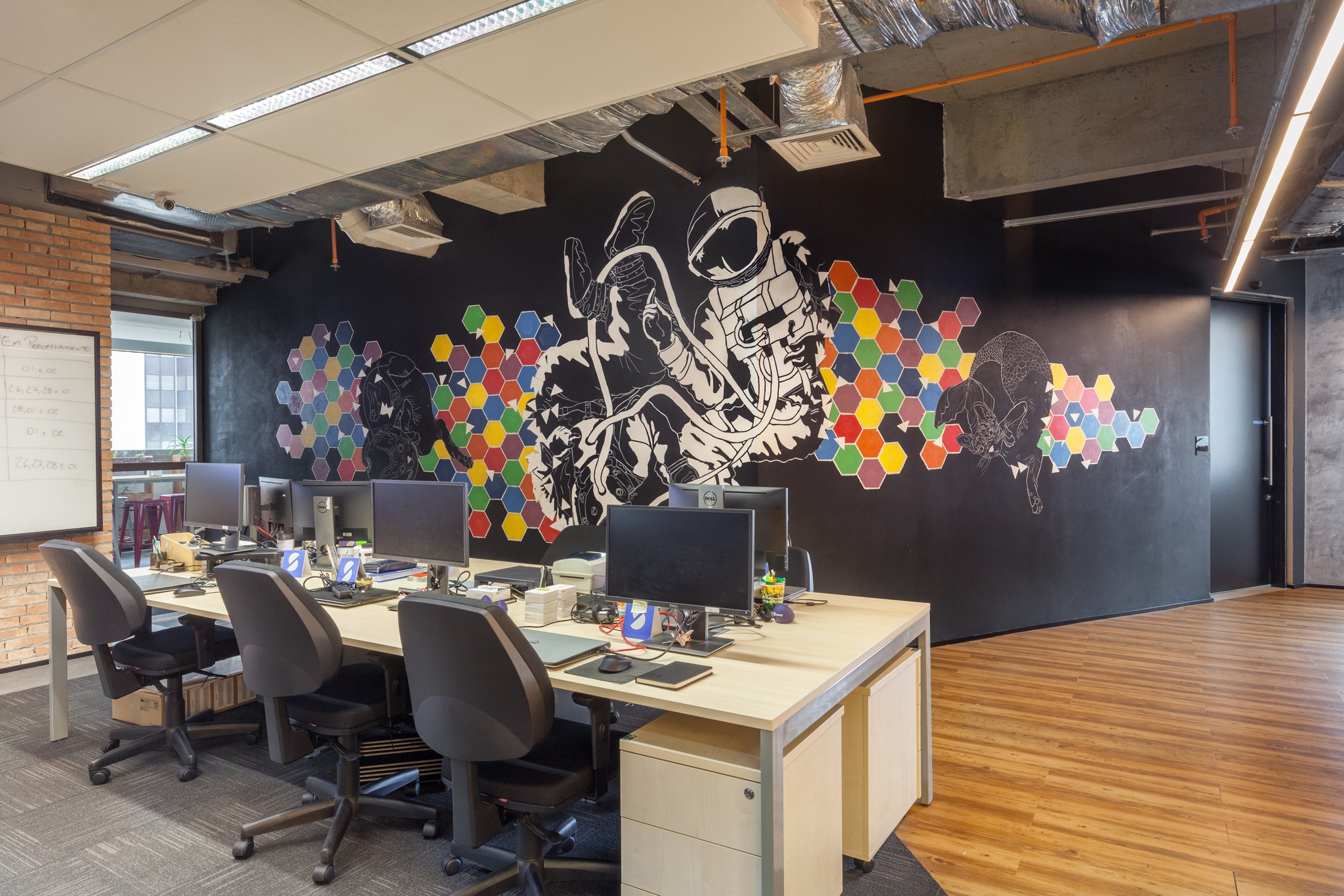 Workstation desks in front of a black wall mural of an astronaut and colorful hexagons, beside an exposed brick wall