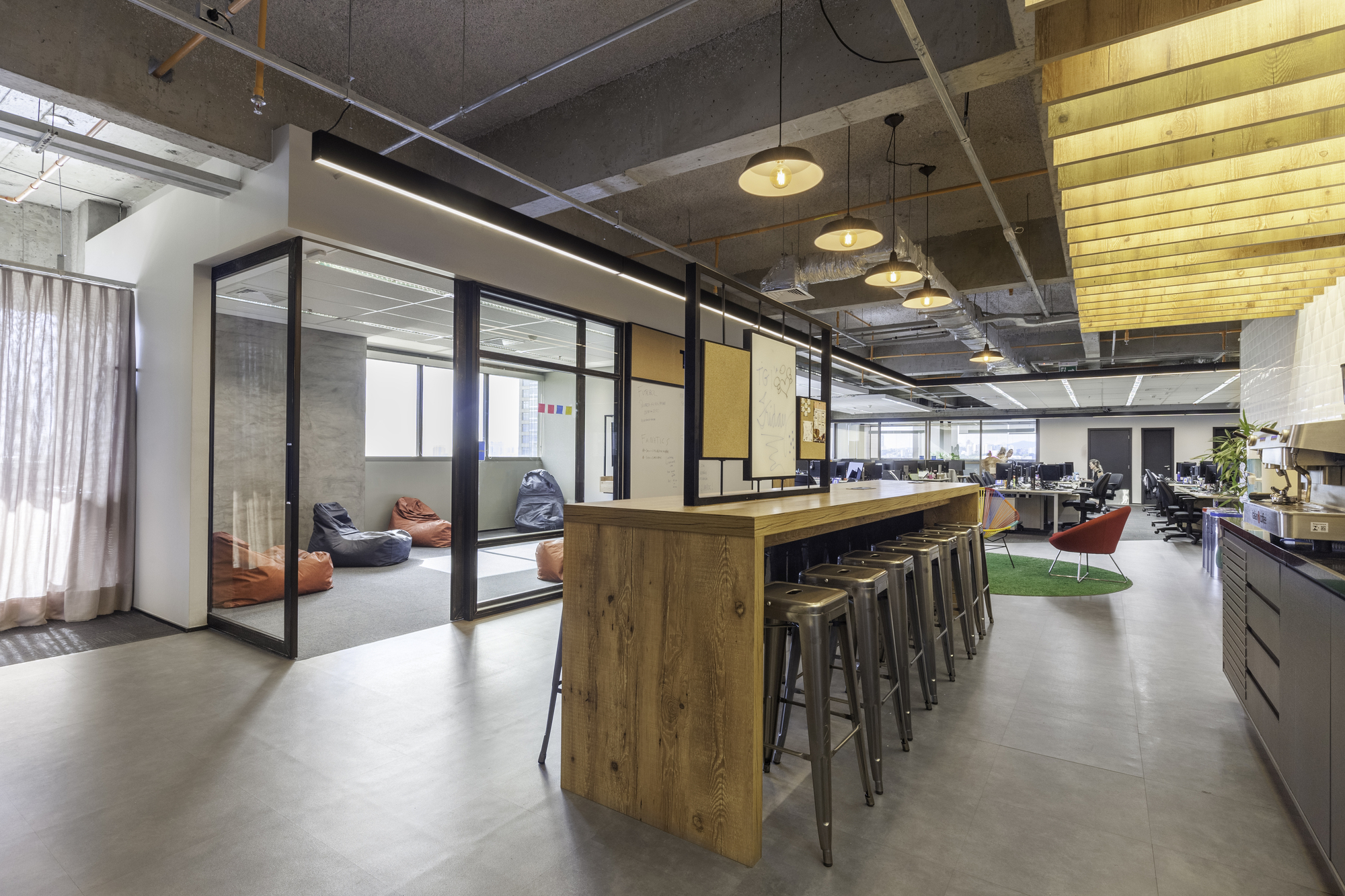 Timber standing-height table with metal stools beside glass-walled meeting room with bean bags, under exposed concrete ceiling and pendant lamps
