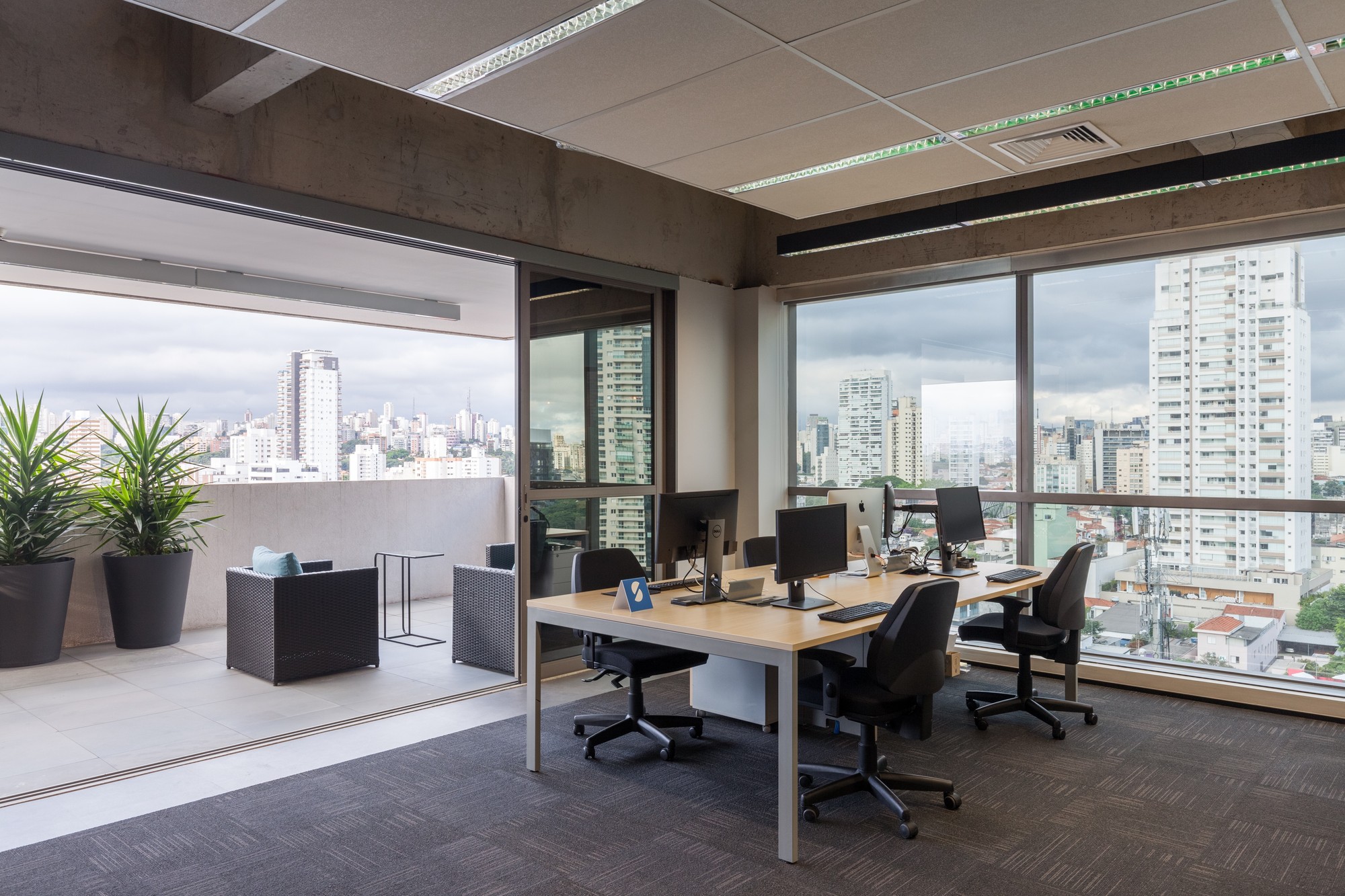 Desk cluster with monitors beside sliding glass door opening to a terrace with potted plants and a hazy city skyline