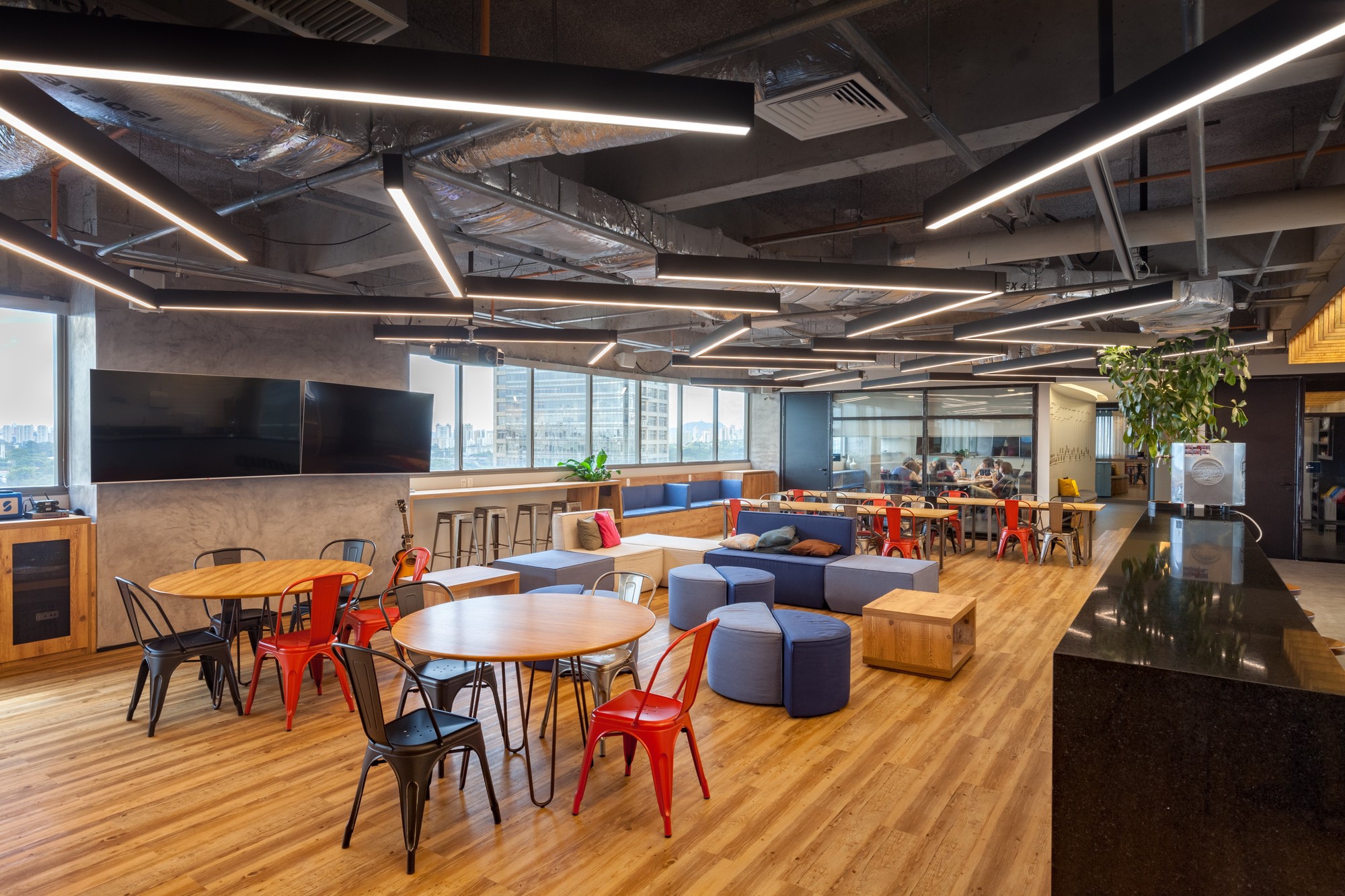 Open lounge with red metal chairs, round tables and blue ottomans beneath black linear light fixtures and exposed ductwork