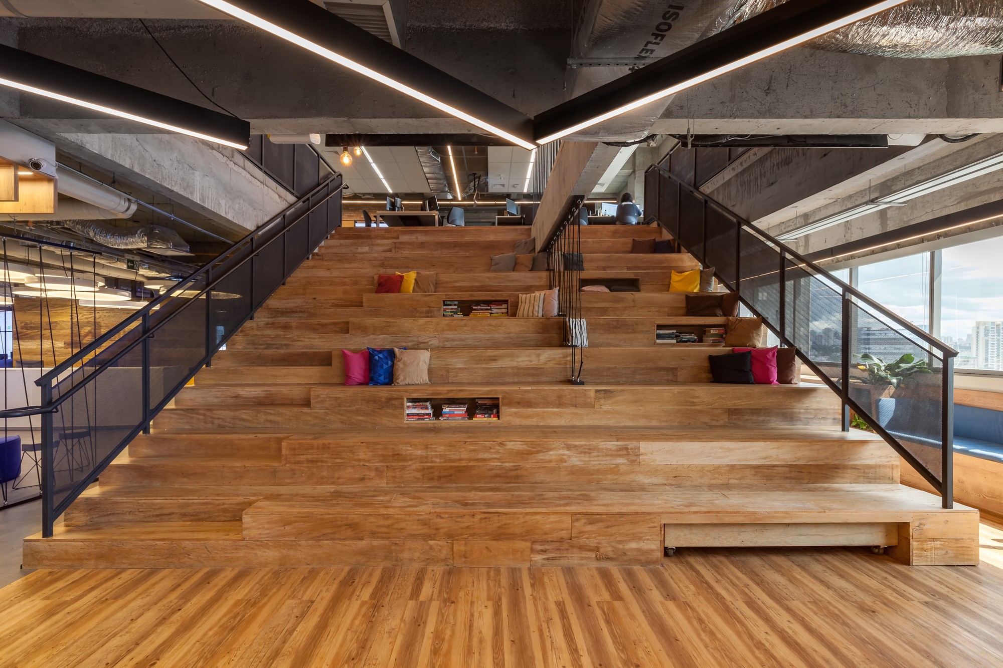 Wide timber stepped seating with colorful cushions and built-in book niches, flanked by glass-panel railings and a window wall