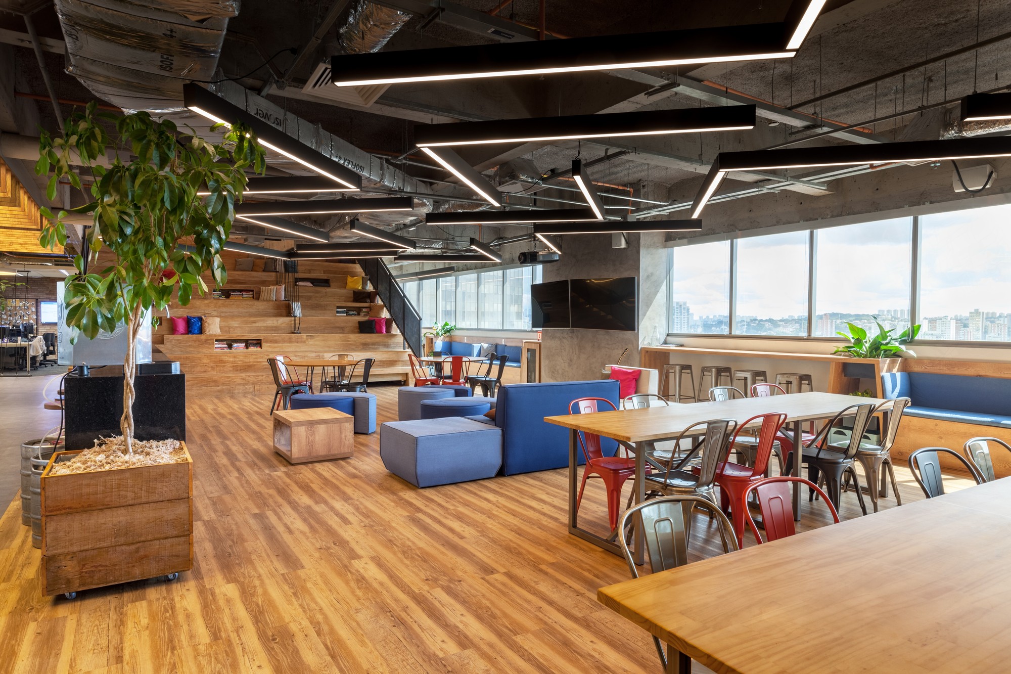 Communal dining area with long wooden tables, red chairs and a potted tree, with stepped seating visible beyond the windows