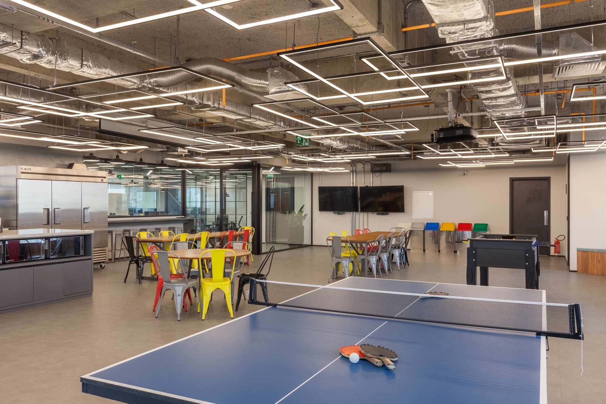 Break room with a blue ping-pong table in the foreground, yellow chairs behind it and square light fixtures under exposed ducts