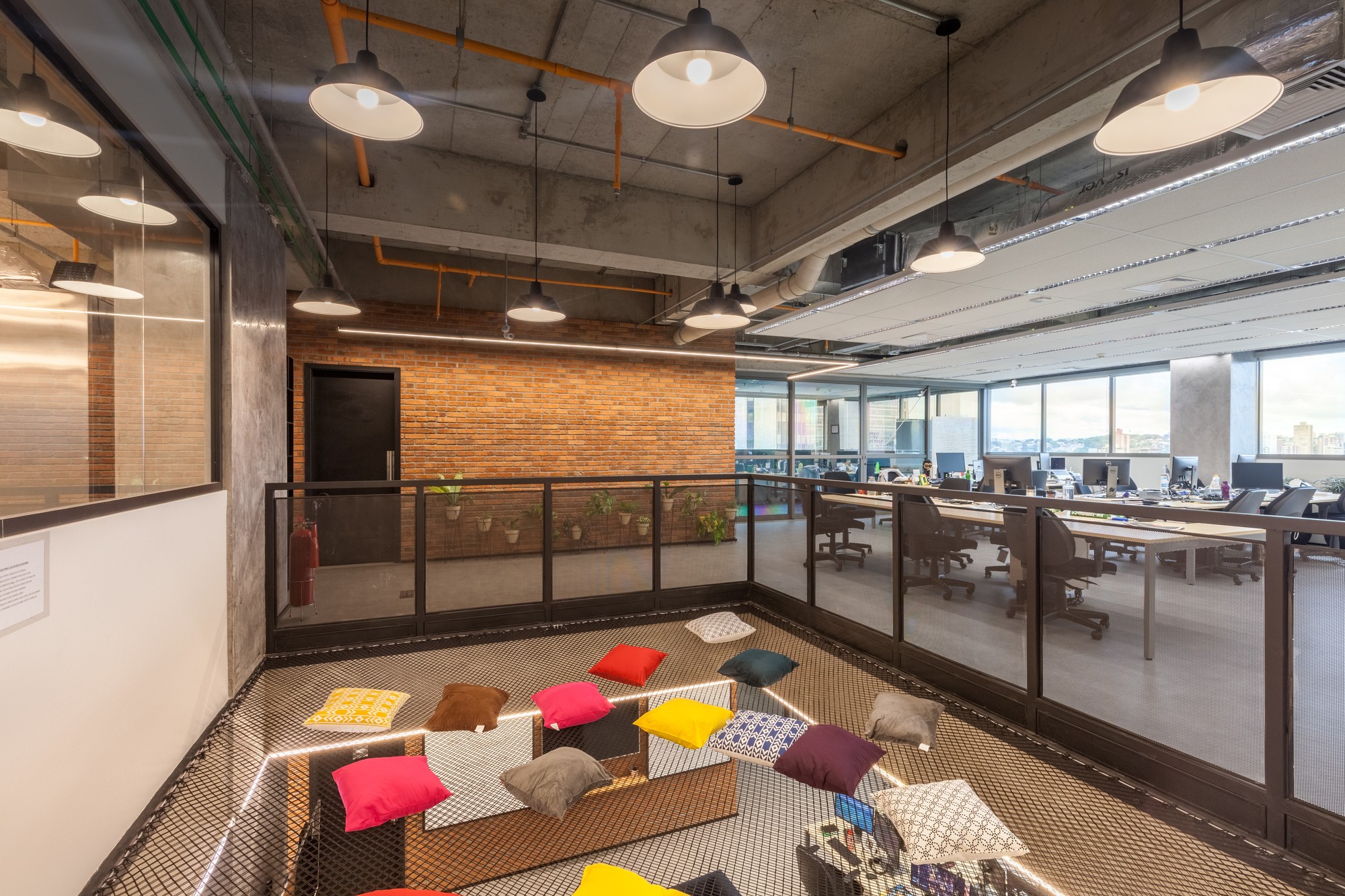Mesh-floored mezzanine scattered with colorful cushions beside a brick wall and glazed railing overlooking a desk area in daylight