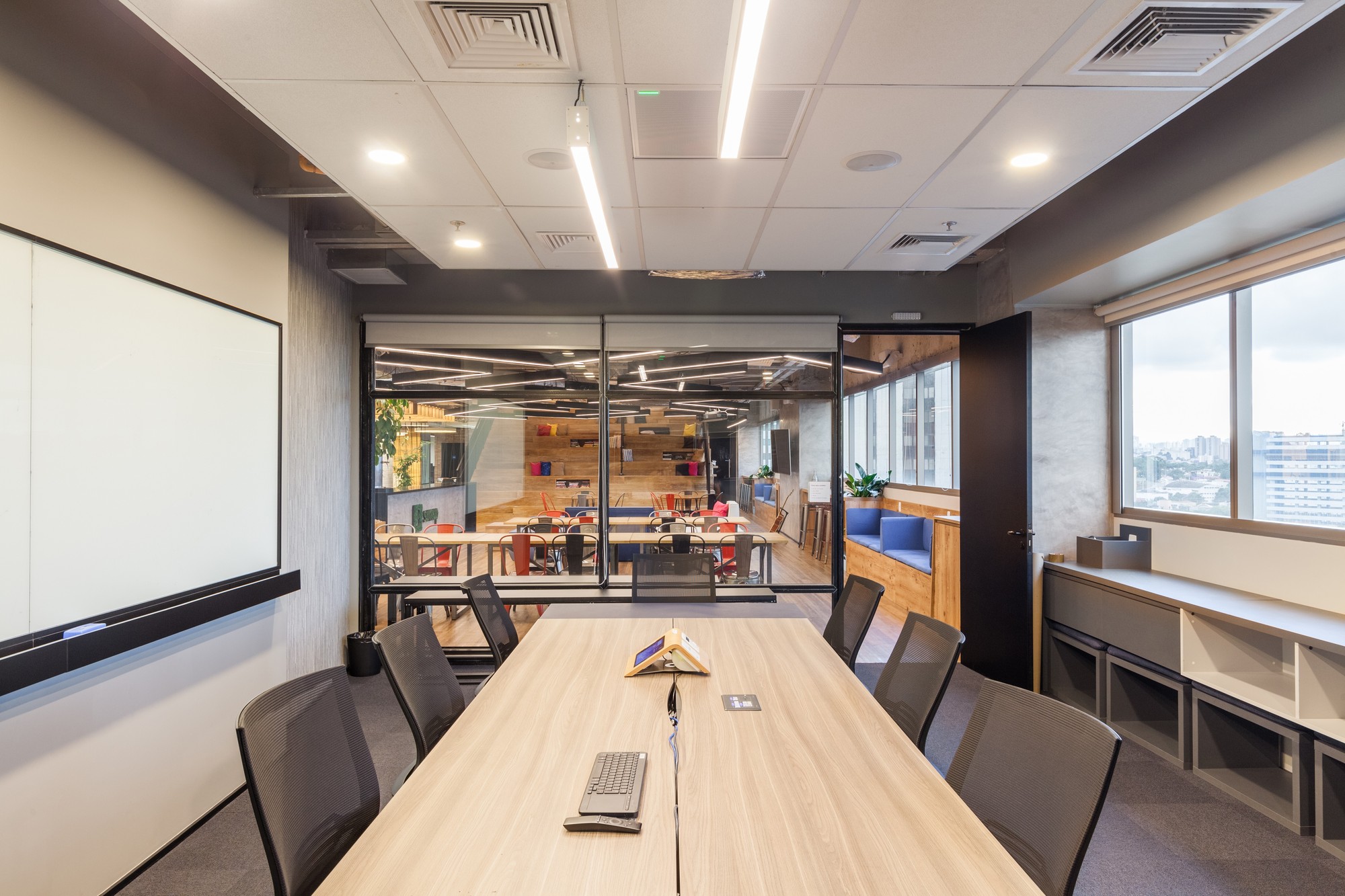Meeting room with a long light wood table, mesh chairs and a whiteboard, with a glass wall opening onto the office and a city view