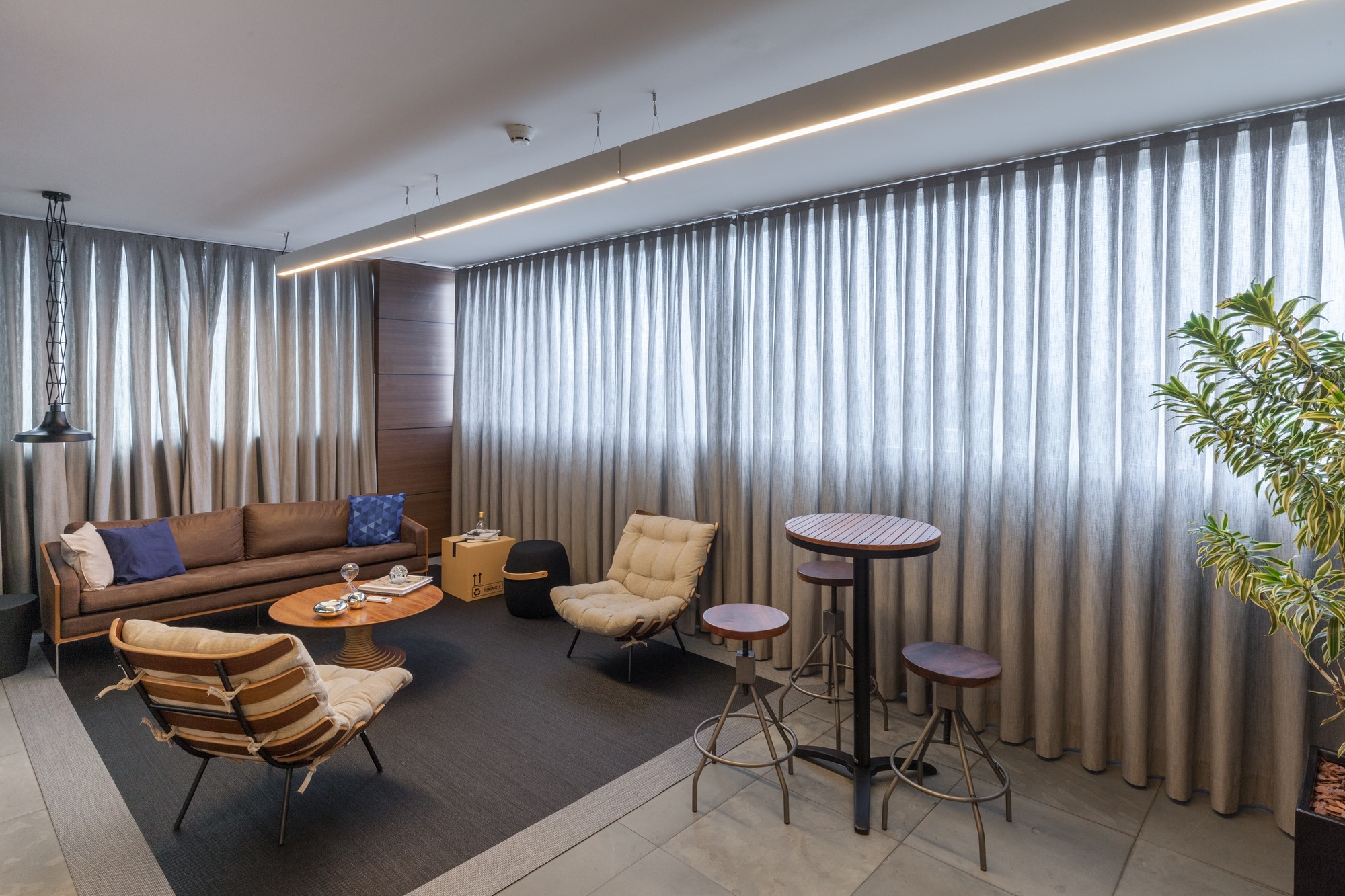 Lounge corner with a brown leather sofa, cane armchairs and high stools, set before floor-length gray curtains and a linear light