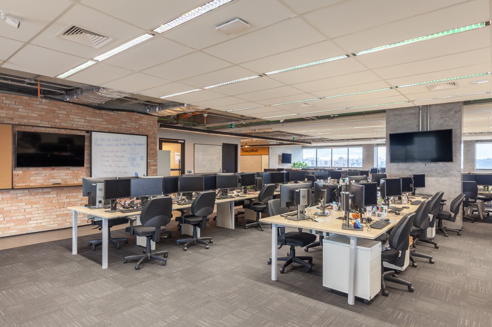 Open office floor with rows of shared desks, monitors and black task chairs beside an exposed brick wall and wall-mounted screen