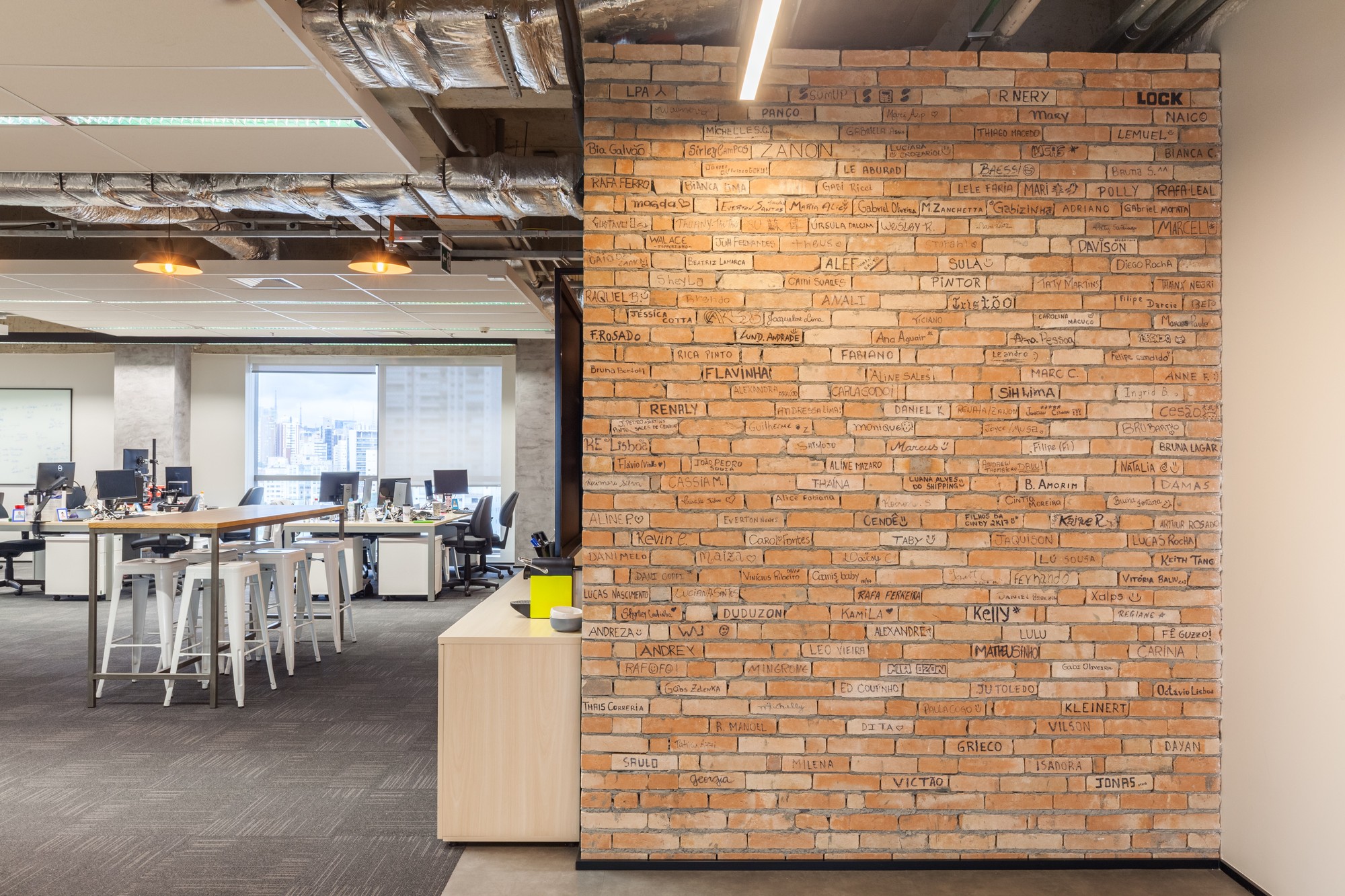 Exposed brick wall covered in handwritten names beside a standing-height table with white stools and a city view through the window