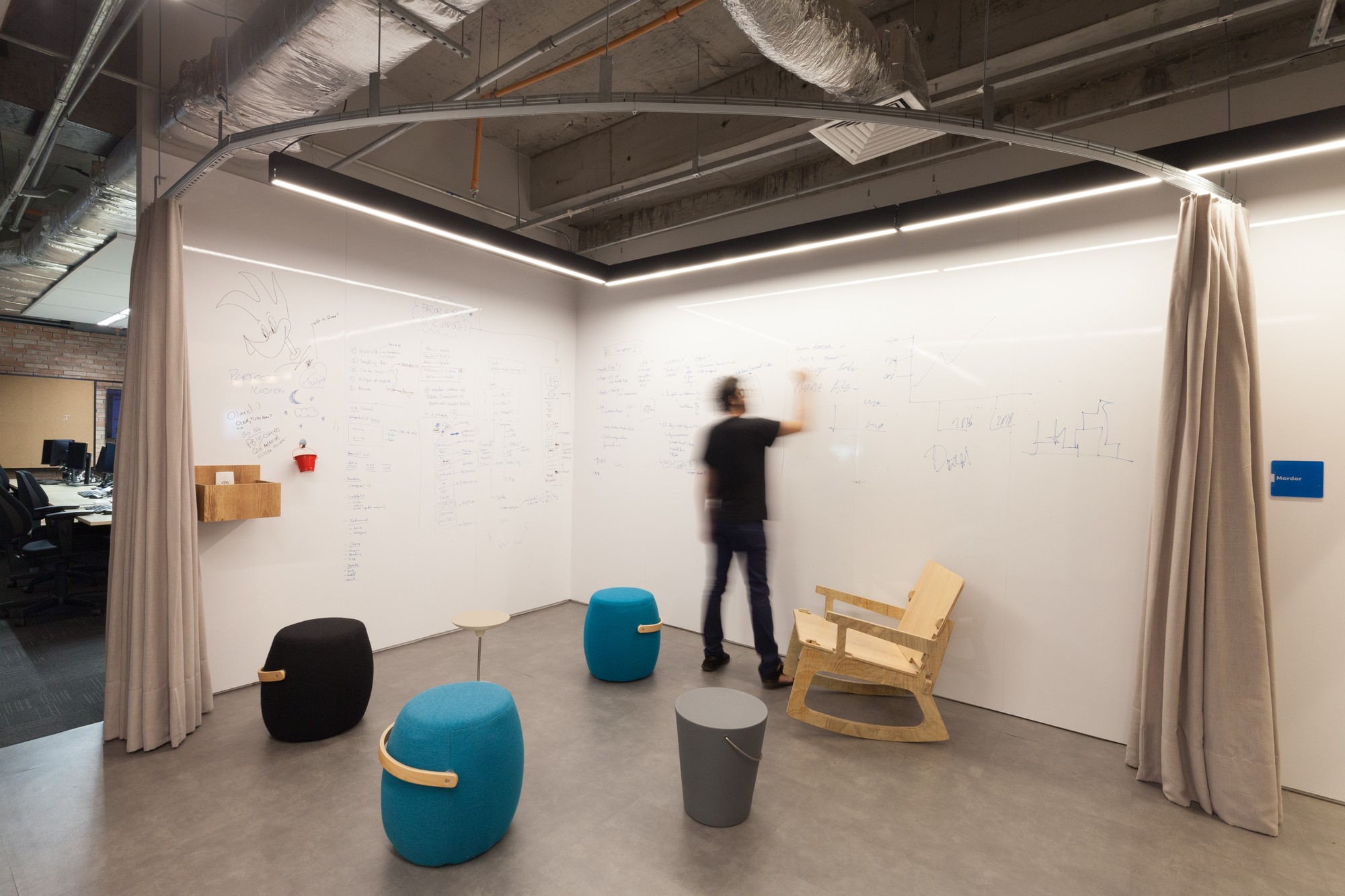 Whiteboard-walled corner with a person writing, colorful stools, a wooden rocking chair and hanging curtains under an exposed ceiling