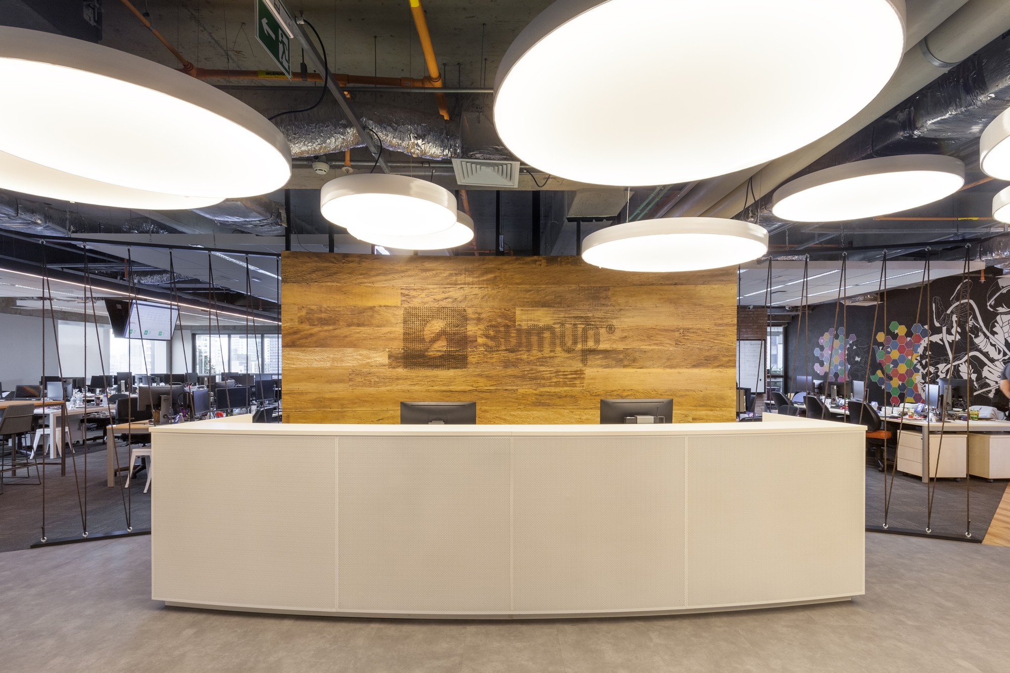 Curved white reception desk in front of a wood-clad logo wall, beneath large round pendant lights and an exposed services ceiling