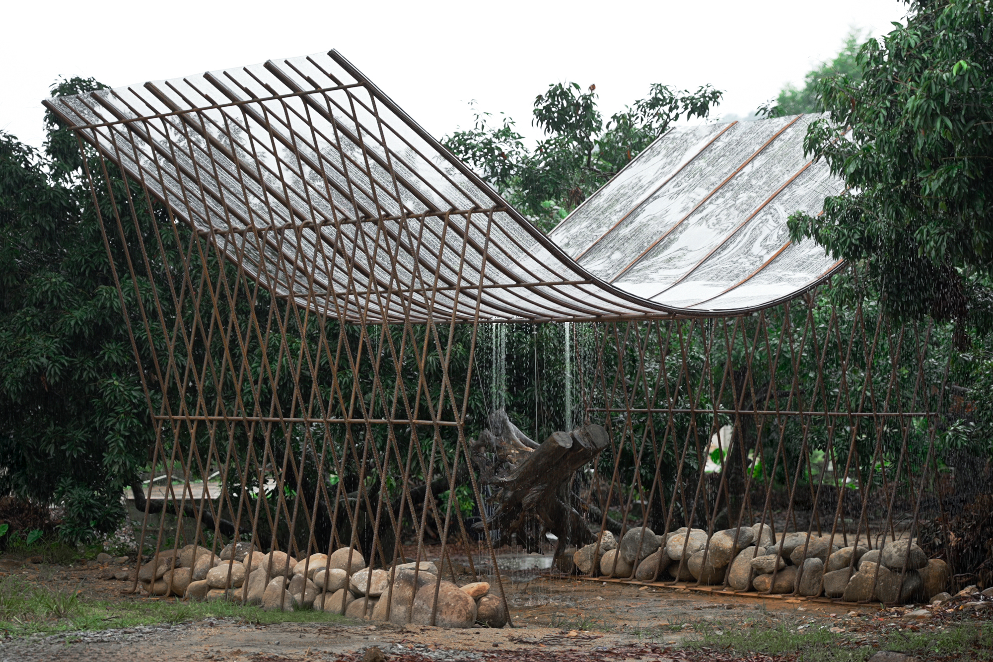 Curved translucent roof held on thin rusted steel rods above stacked river stones and a driftwood stump, with rain falling through the orchard