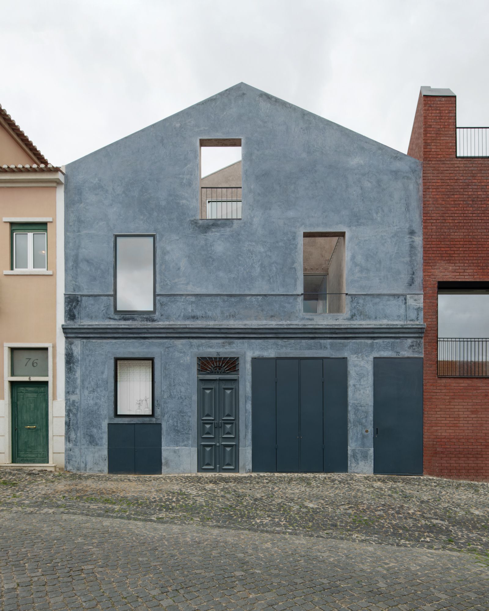 Street facade of blue-grey plastered gabled house with cut-out openings and dark panel doors, set between a pink neighbour and red brick under overcast sky
