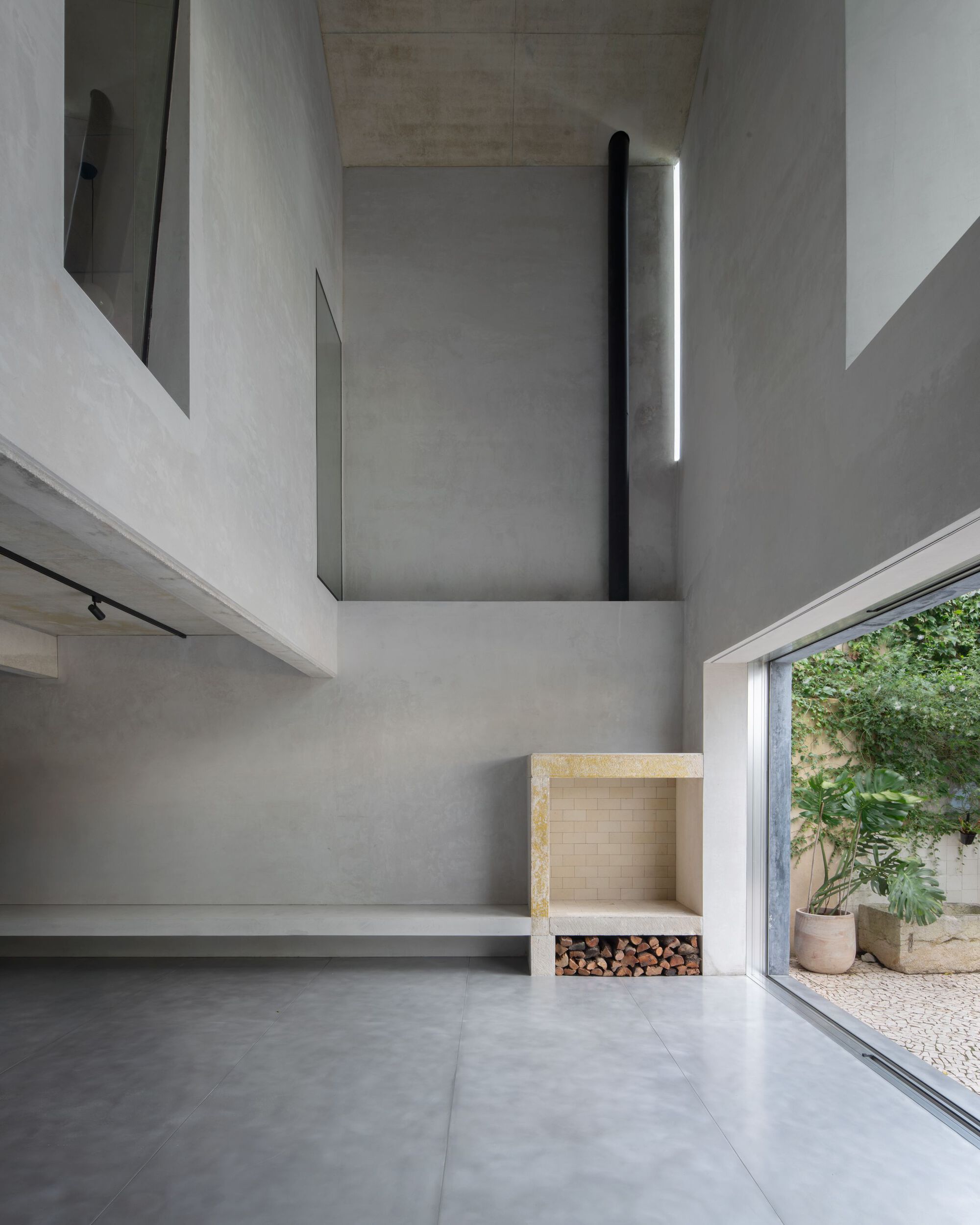Double-height concrete living space with a stone fireplace and log storage, opening through a sliding glass wall to a planted gravel courtyard