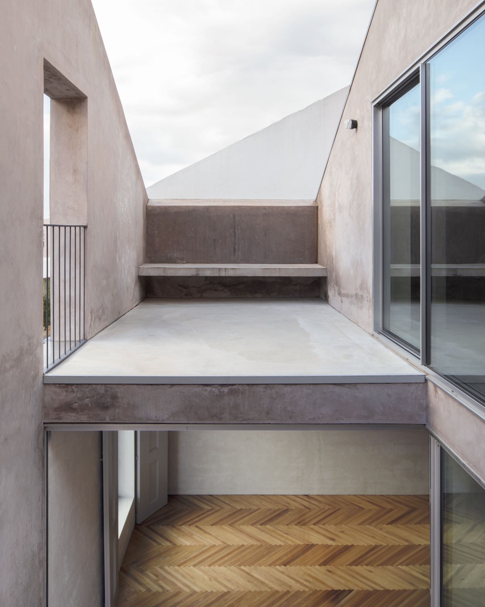 Narrow roof terrace between pitched concrete walls with a built-in ledge, a railing and herringbone timber floor visible in the room below