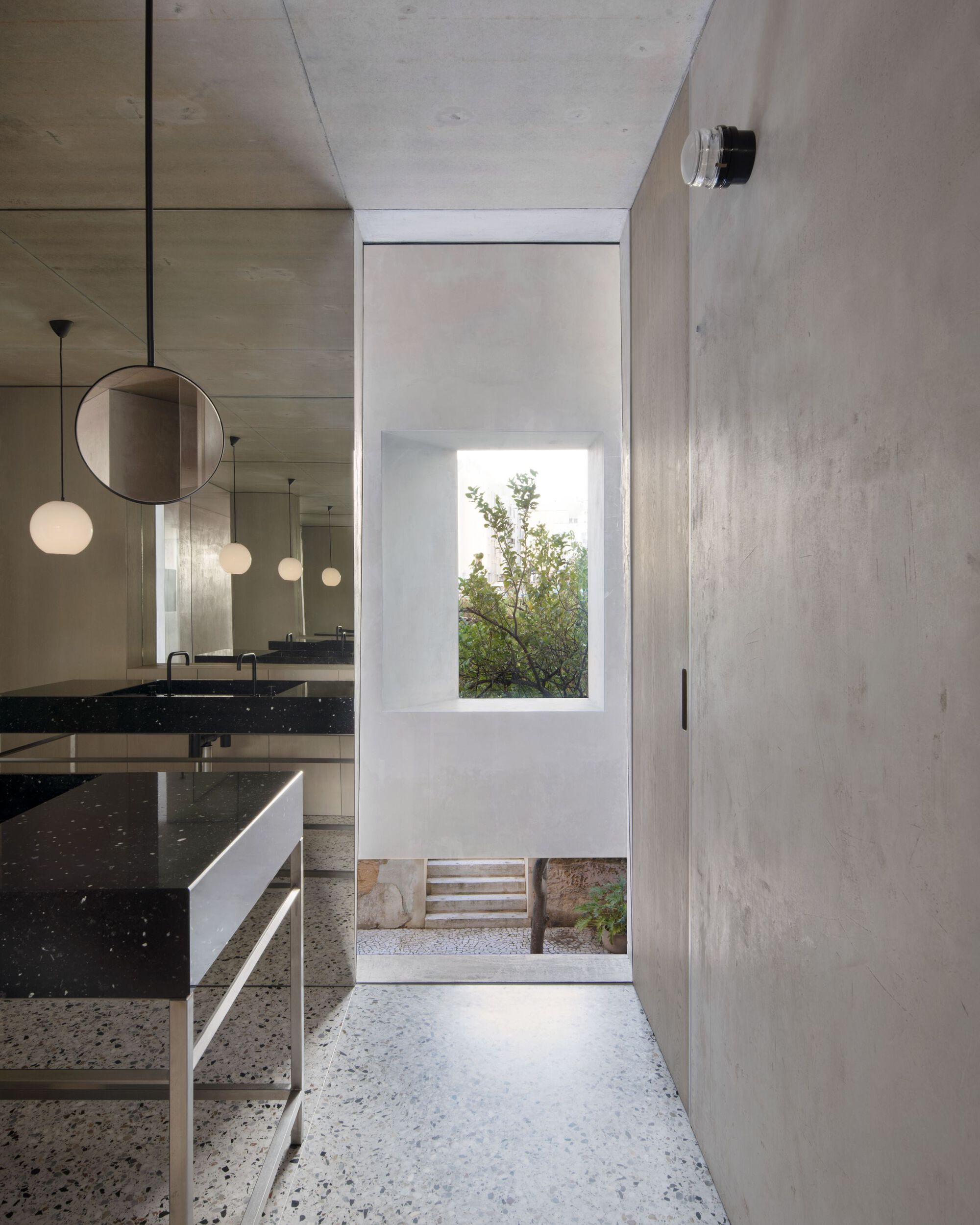 Bathroom corridor with terrazzo floor, black stone vanity and round mirror, framing a window onto a garden tree in daylight