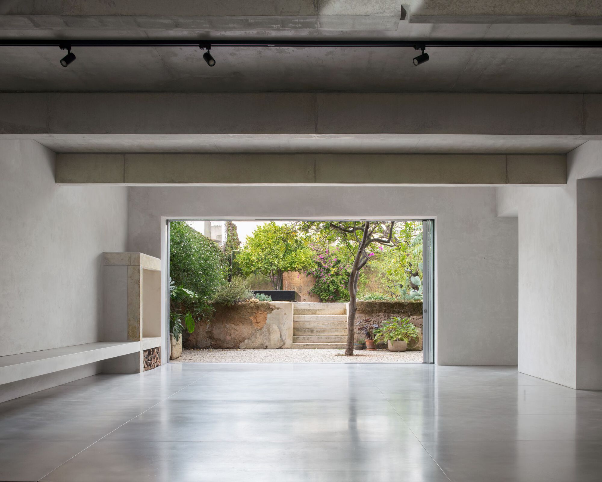 Open concrete-ceilinged room with polished floor and built-in bench, looking through a wide opening to garden steps and a citrus tree in daylight