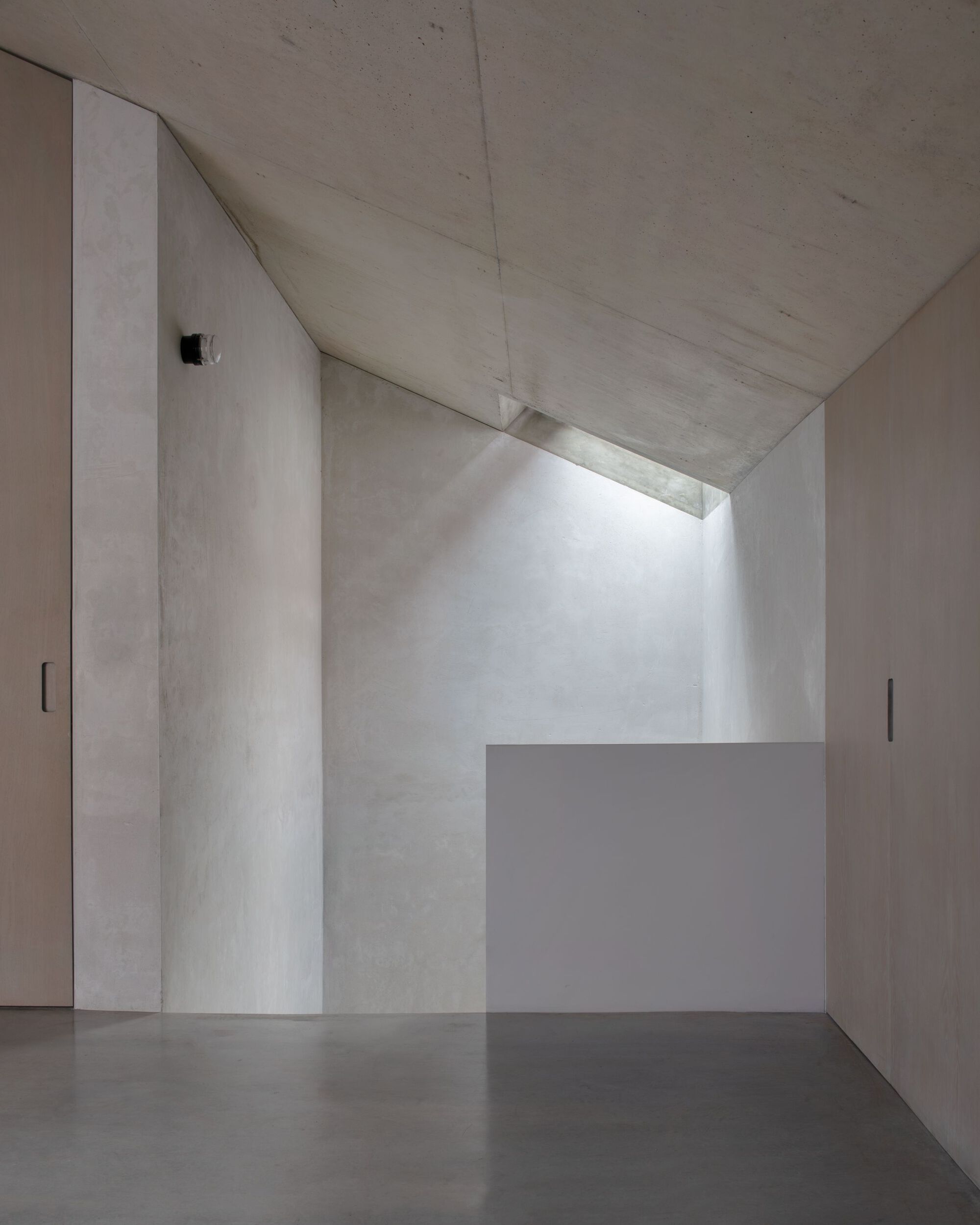 Sloping concrete ceiling with a narrow skylight washing light down grey walls, beside pale timber doors and a low white parapet