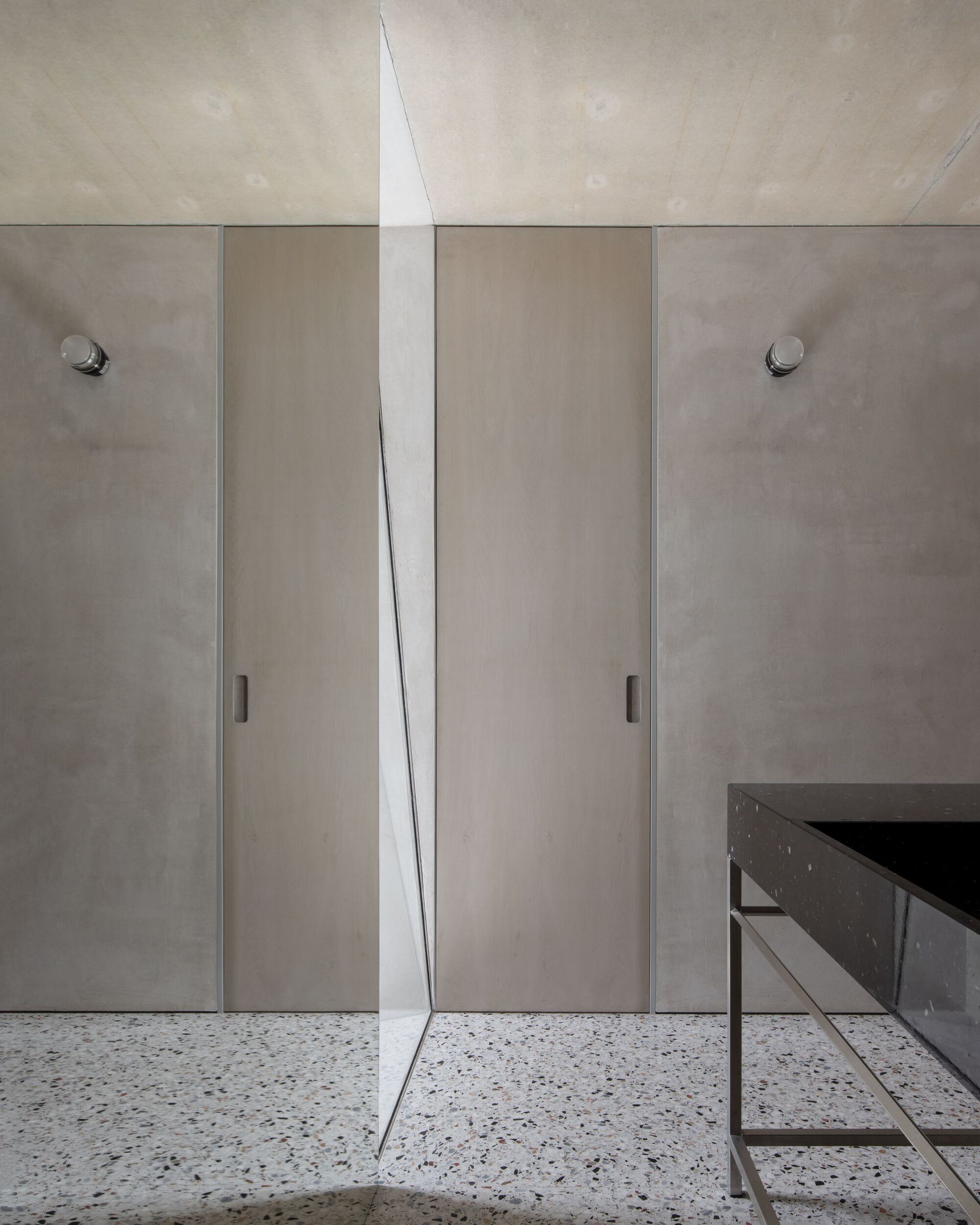 Two tall pale timber doors set between concrete-toned wall panels, with a mirrored edge, terrazzo floor and dark stone basin in soft light