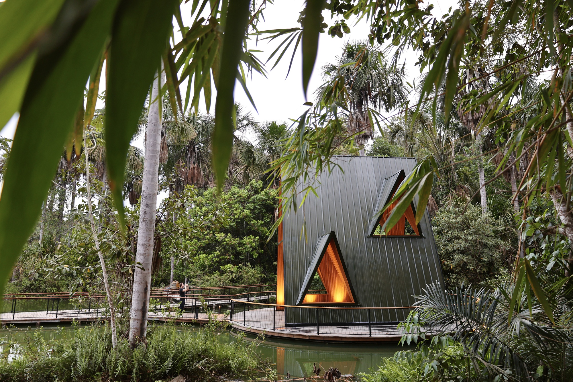 A-frame pavilion clad in dark standing-seam metal with glowing triangular openings, seen through bamboo leaves beside a pond on an overcast day