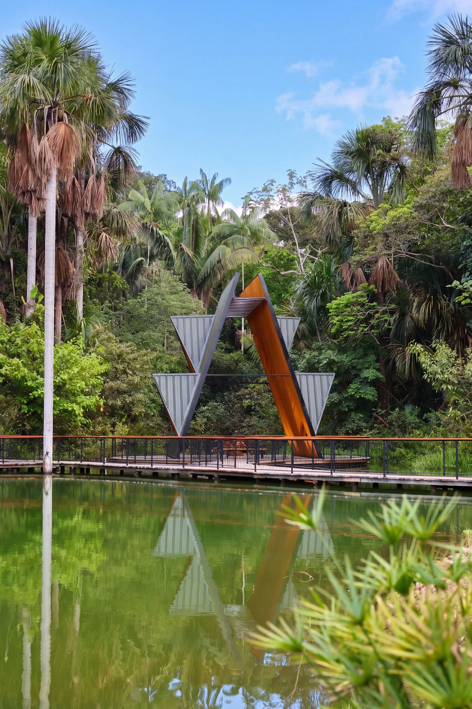 Pavilion's inverted V frame of steel and timber reflected in a green pond under a blue sky with palms behind