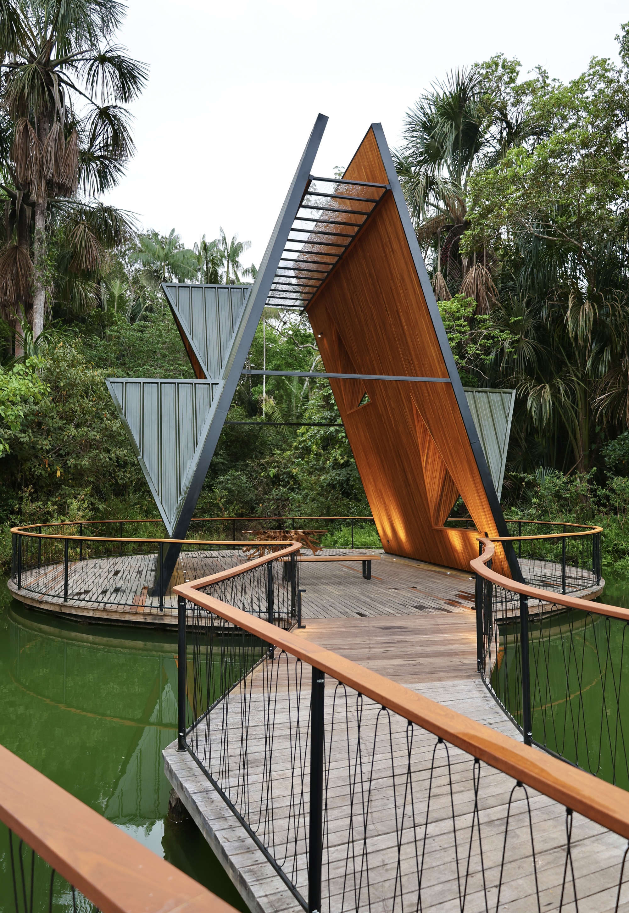Timber boardwalk with cable-mesh railings leading to a circular deck and the angled V-shaped pavilion over green water