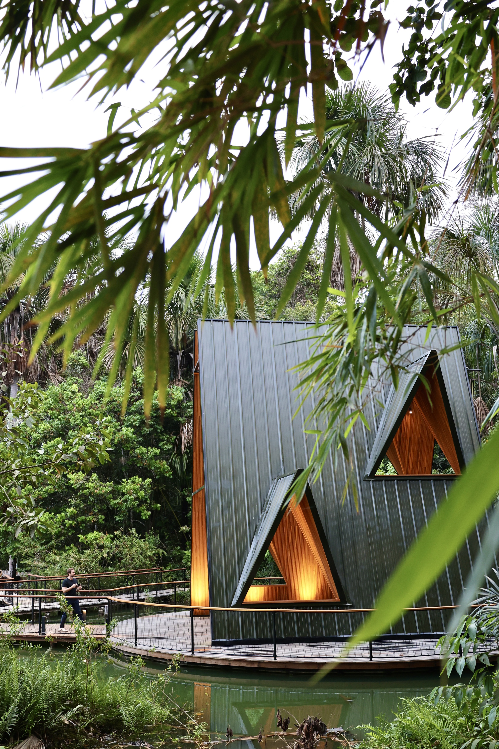 Dark metal pavilion facade with two lit triangular windows, framed by hanging bamboo leaves while a visitor walks the boardwalk